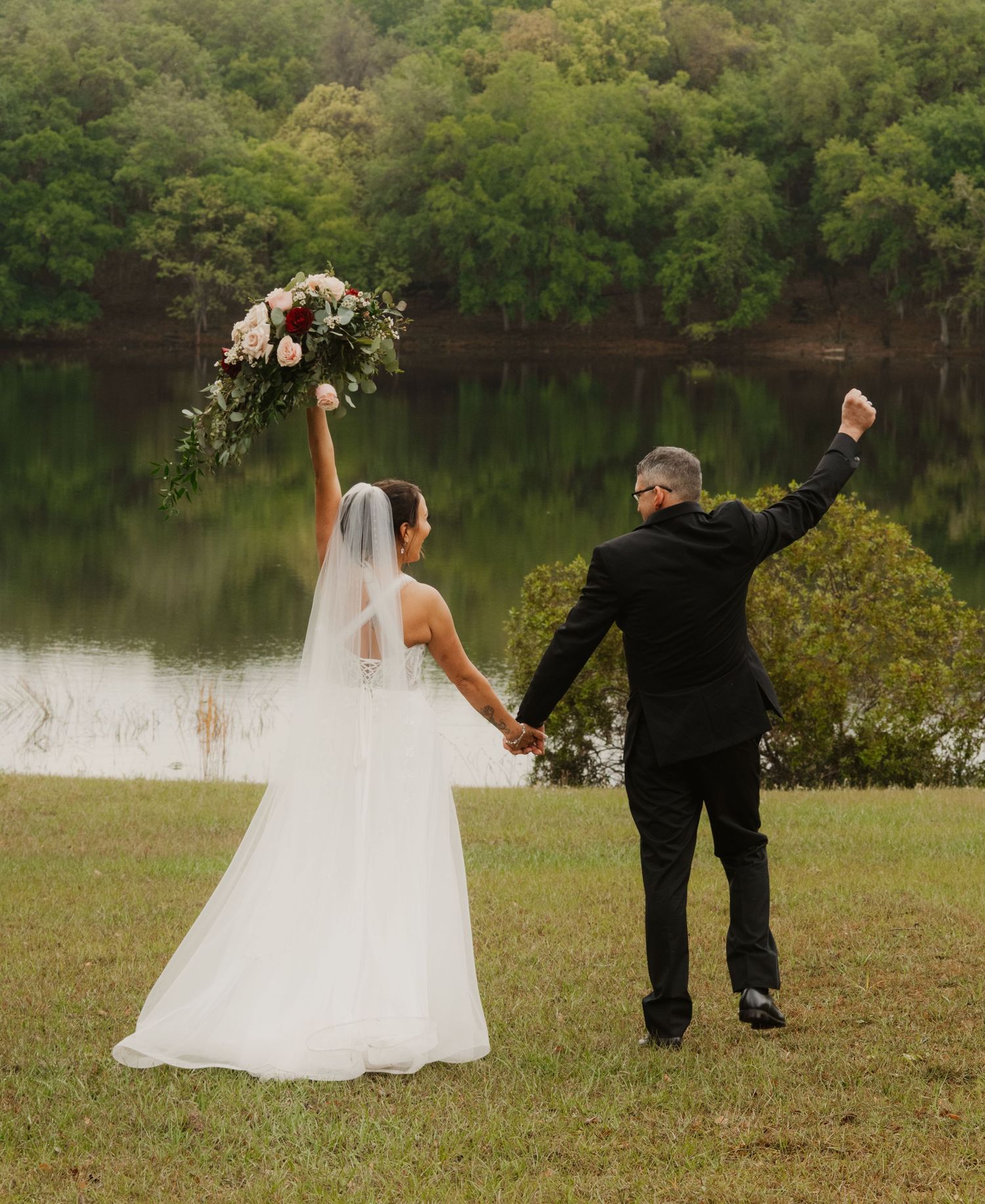 A couple in formal wedding attire dances playfully in a grassy field by a lake at sunset.
