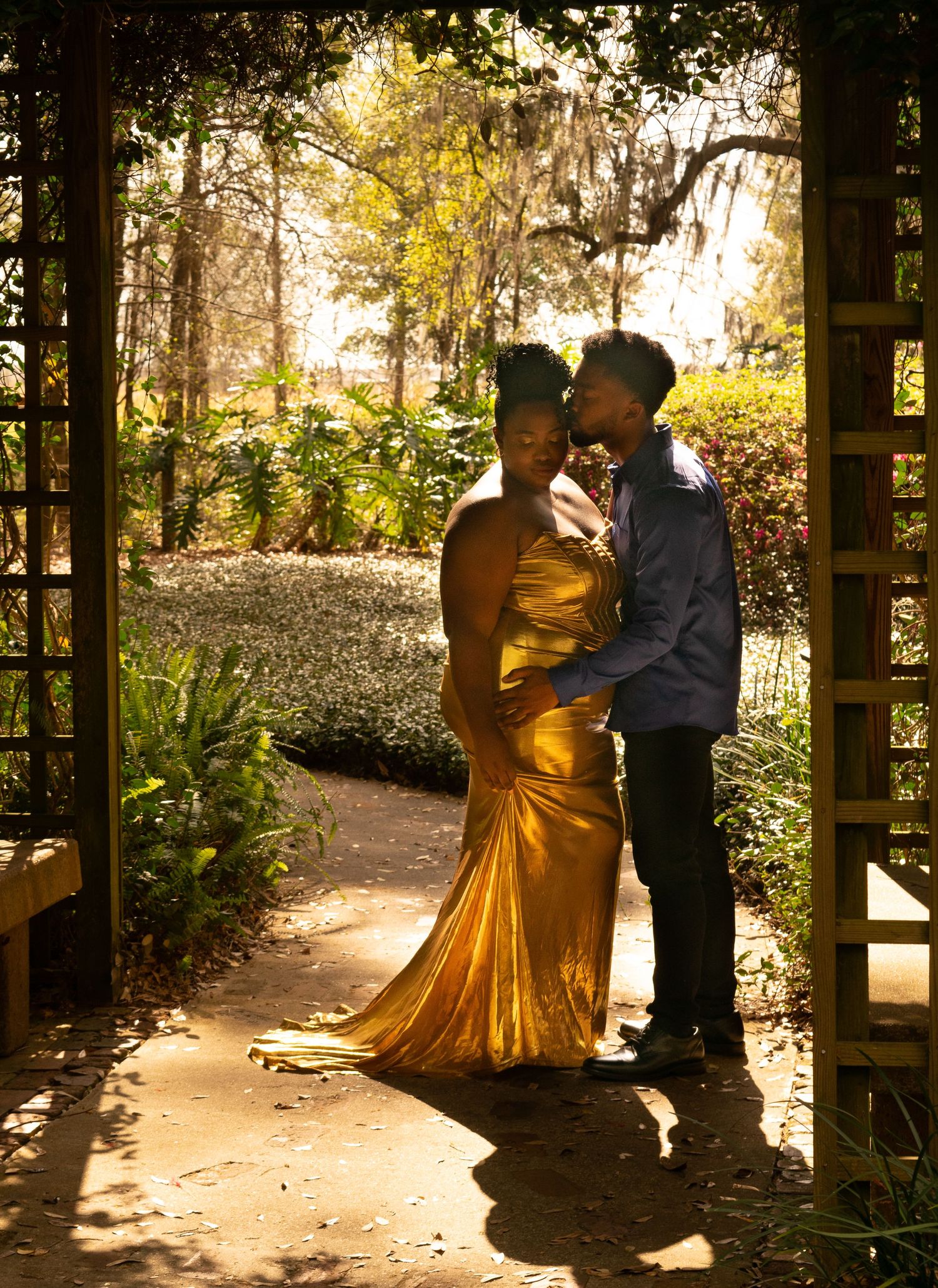 A couple in formal attire shares an intimate dance moment under a wooded canopy with golden sunlight filtering through.