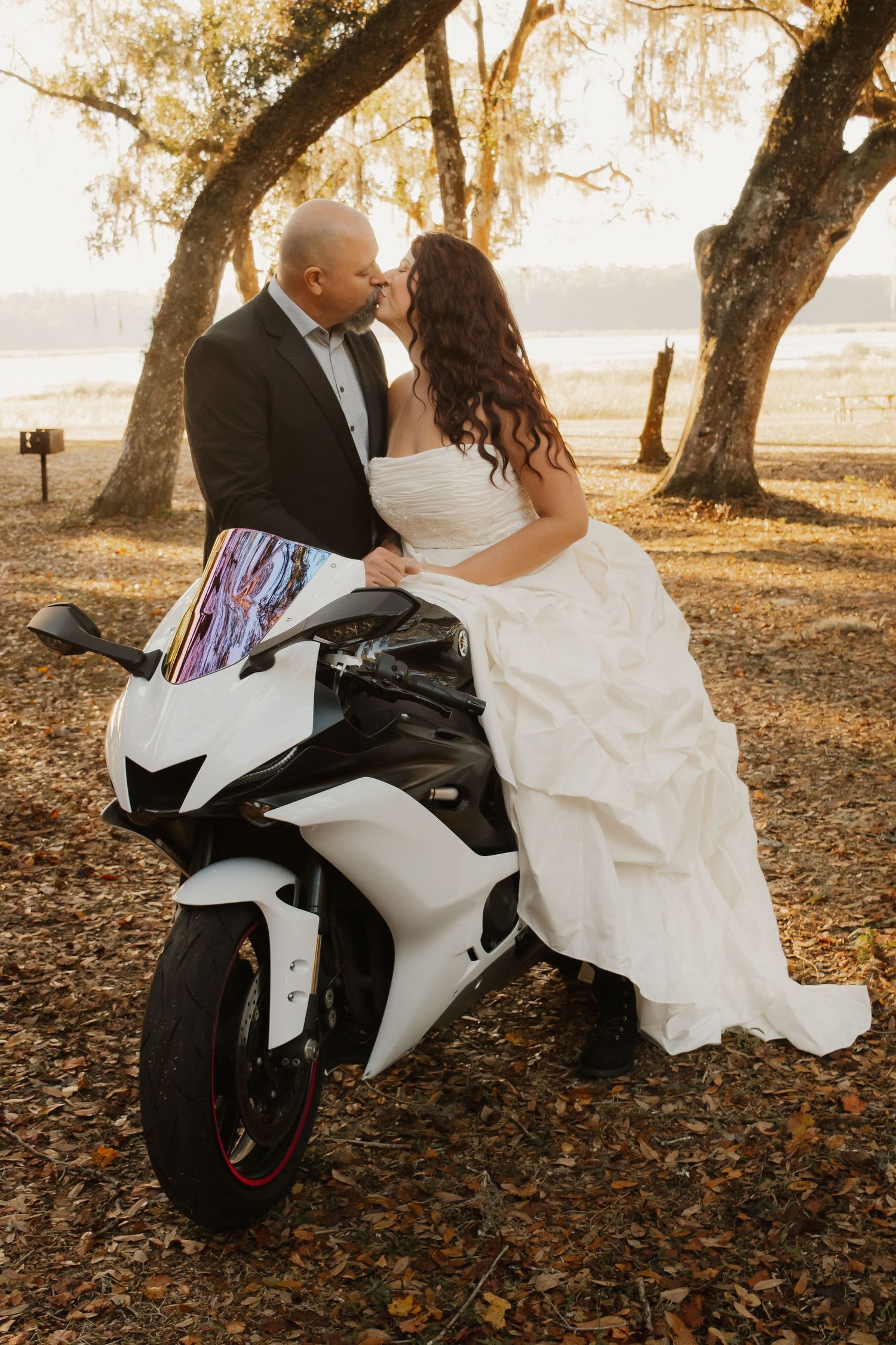 Romantic wedding couple poses with white motorcycle under autumn trees at sunset in a rustic outdoor setting.