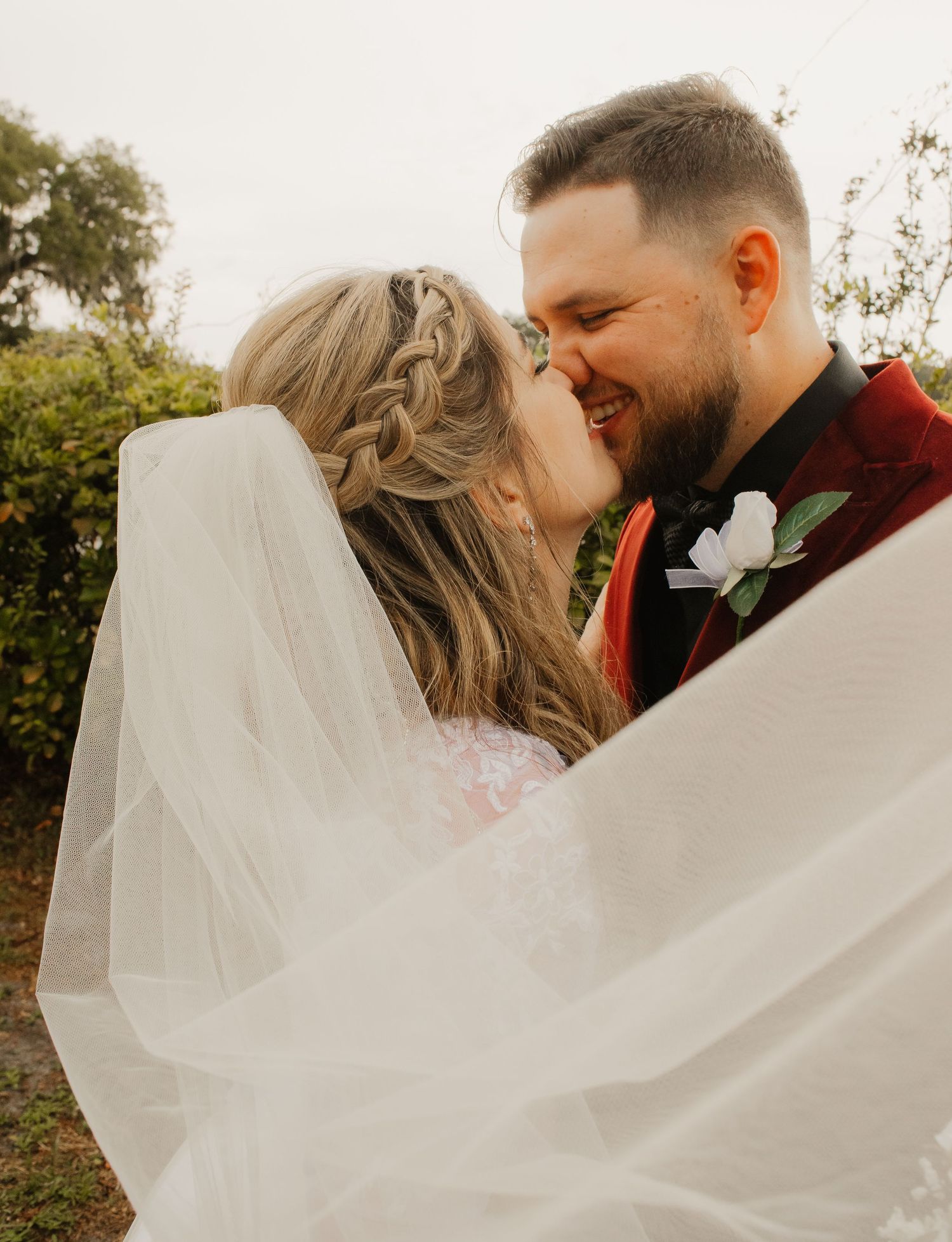 A romantic wedding couple embraces outdoors as the bride's veil flows dramatically in the wind during their photoshoot.
