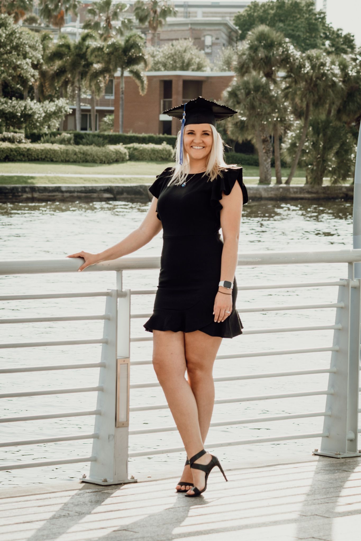 Graduate in black dress and cap poses on white boardwalk near water during sunny outdoor photoshoot.