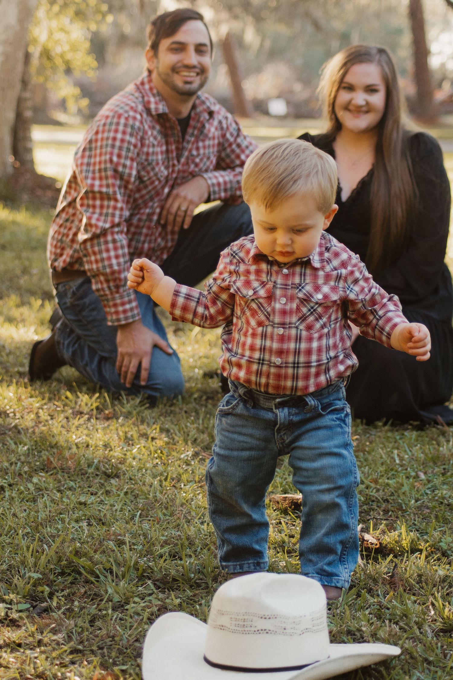A toddler in a plaid shirt and jeans reaches down to touch a white cowboy hat while sitting on grass at sunset.