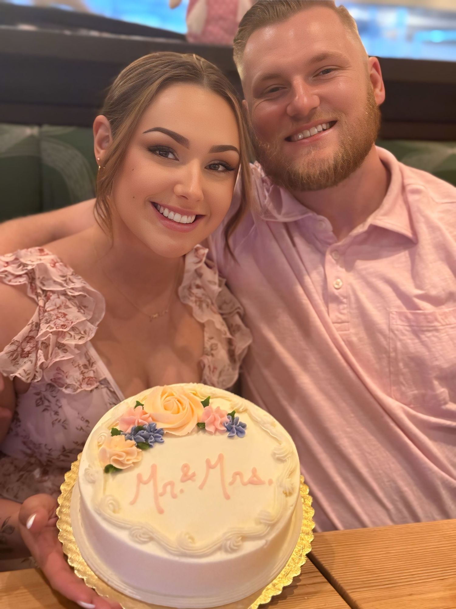 A couple smiles while holding a decorated white cake with yellow flowers and blue accents at their dinner after their wedding day.