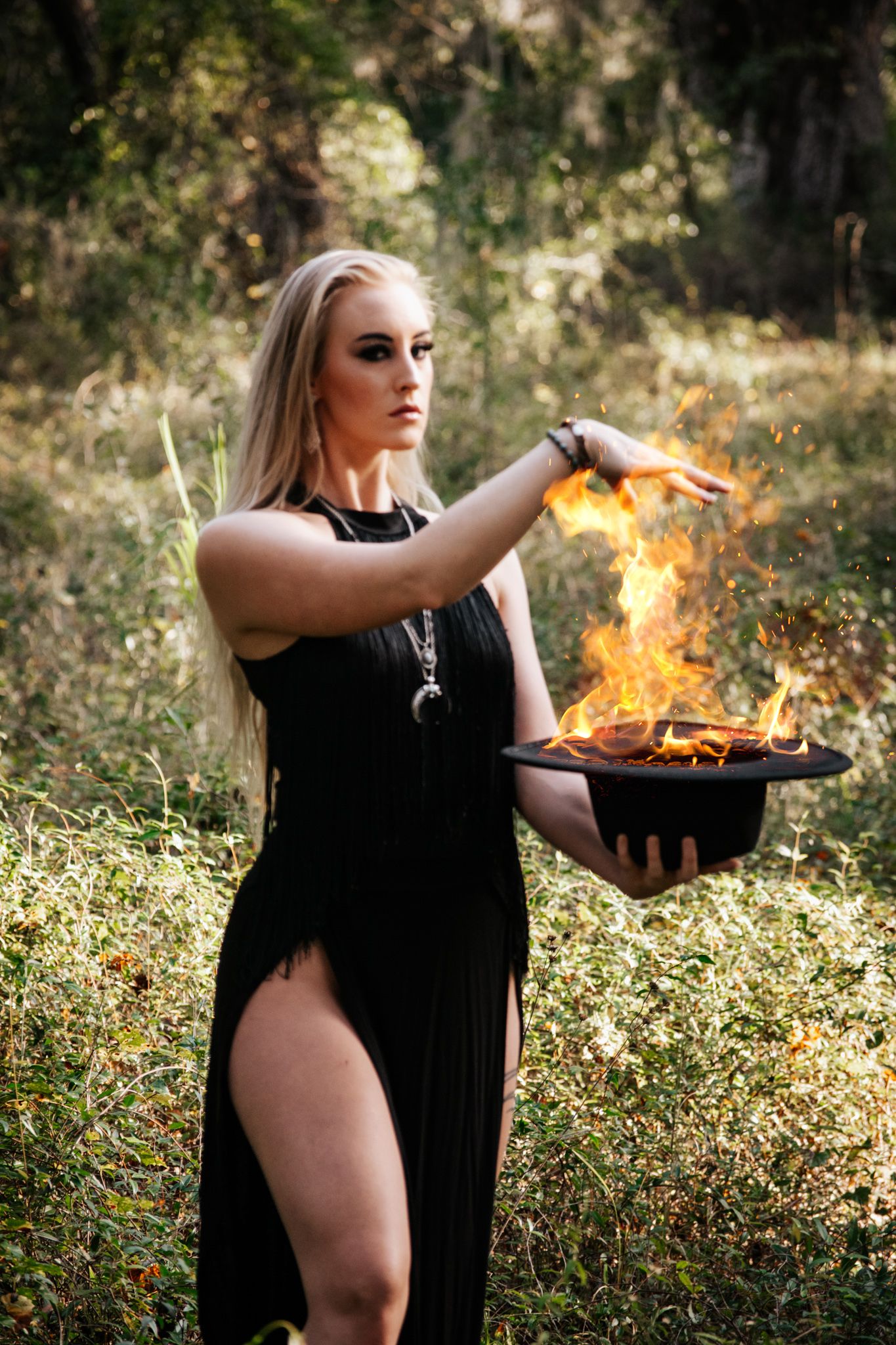 A woman in a black dress performs a mystical ritual with fire in a wooded outdoor setting.