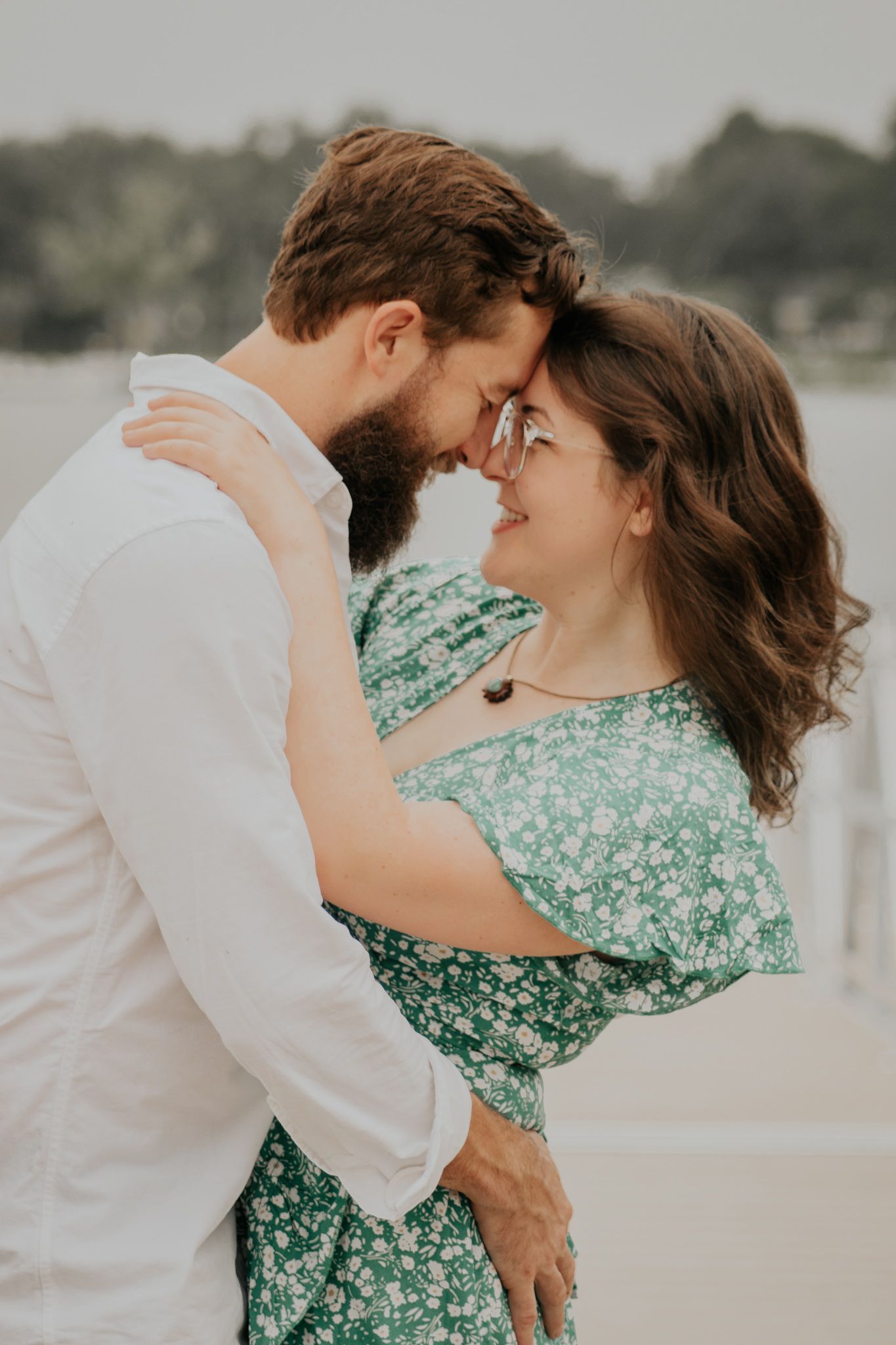 A couple embraces on a windy dock with a lake behind them.