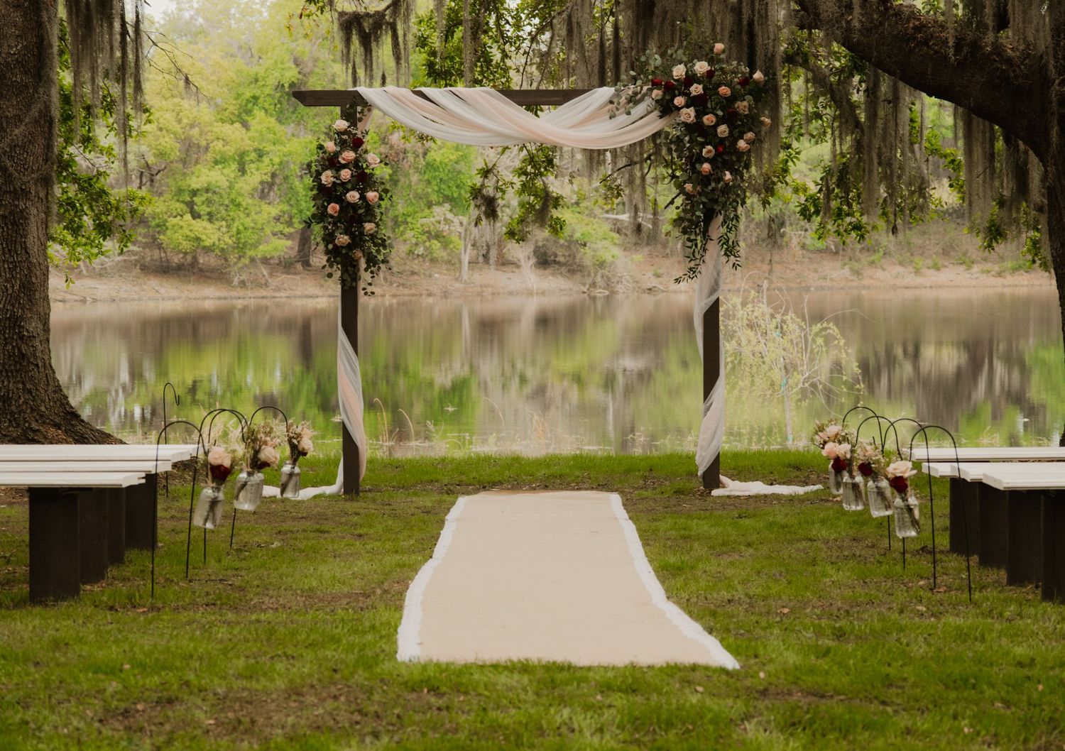 A panoramic view of a serene garden walkway leading to a lake surrounded by moss-draped oak trees and lush foliage.