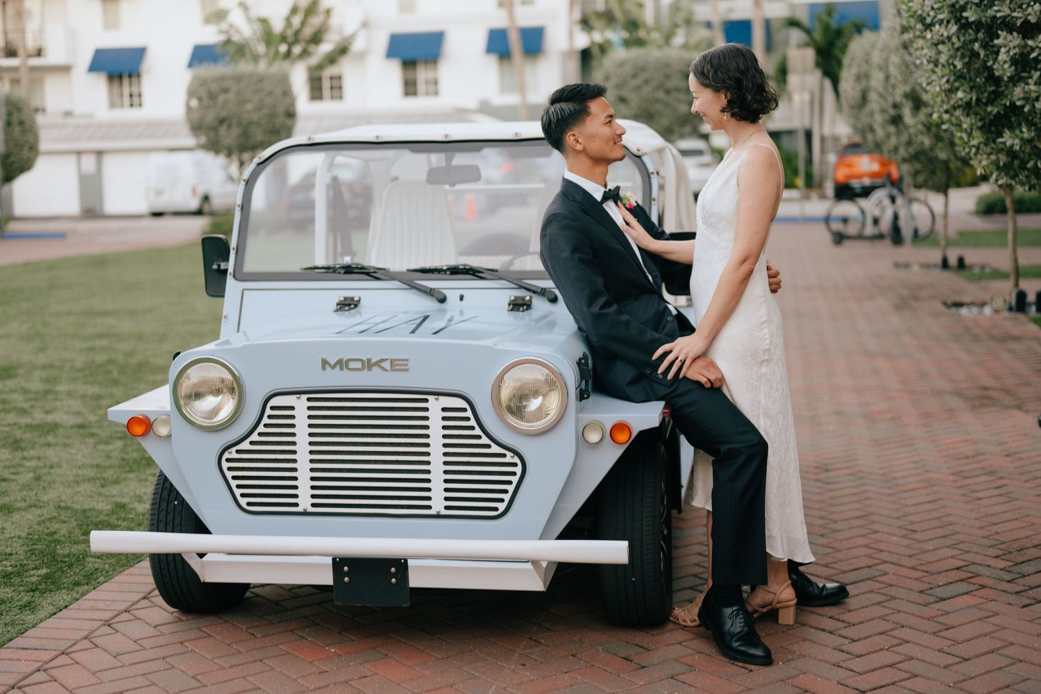 A wedding couple poses together with a vintage light blue Mini car on a brick pathway.