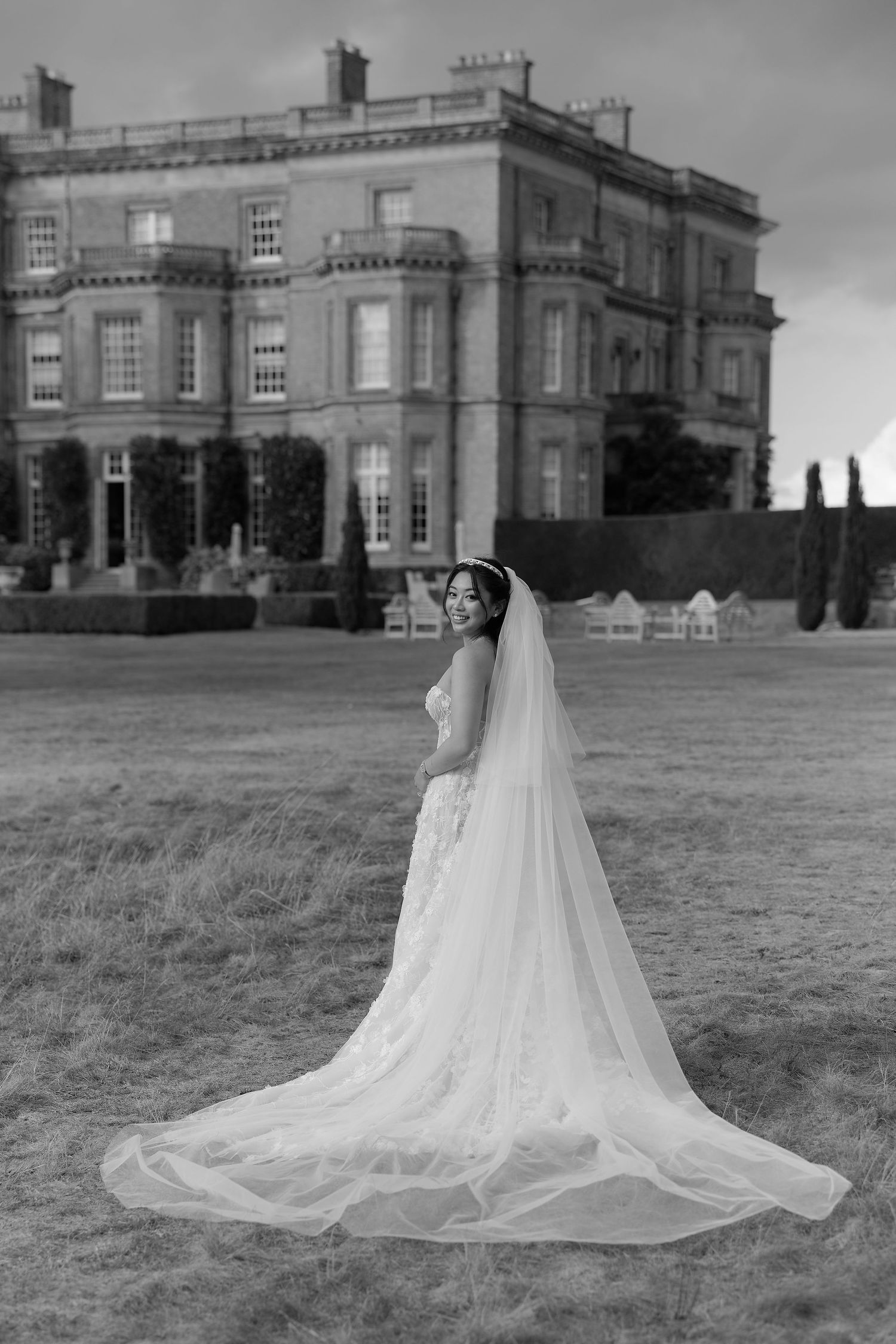 Black and white photo of a bride in a flowing gown and long veil standing on a lawn before a grand estate.