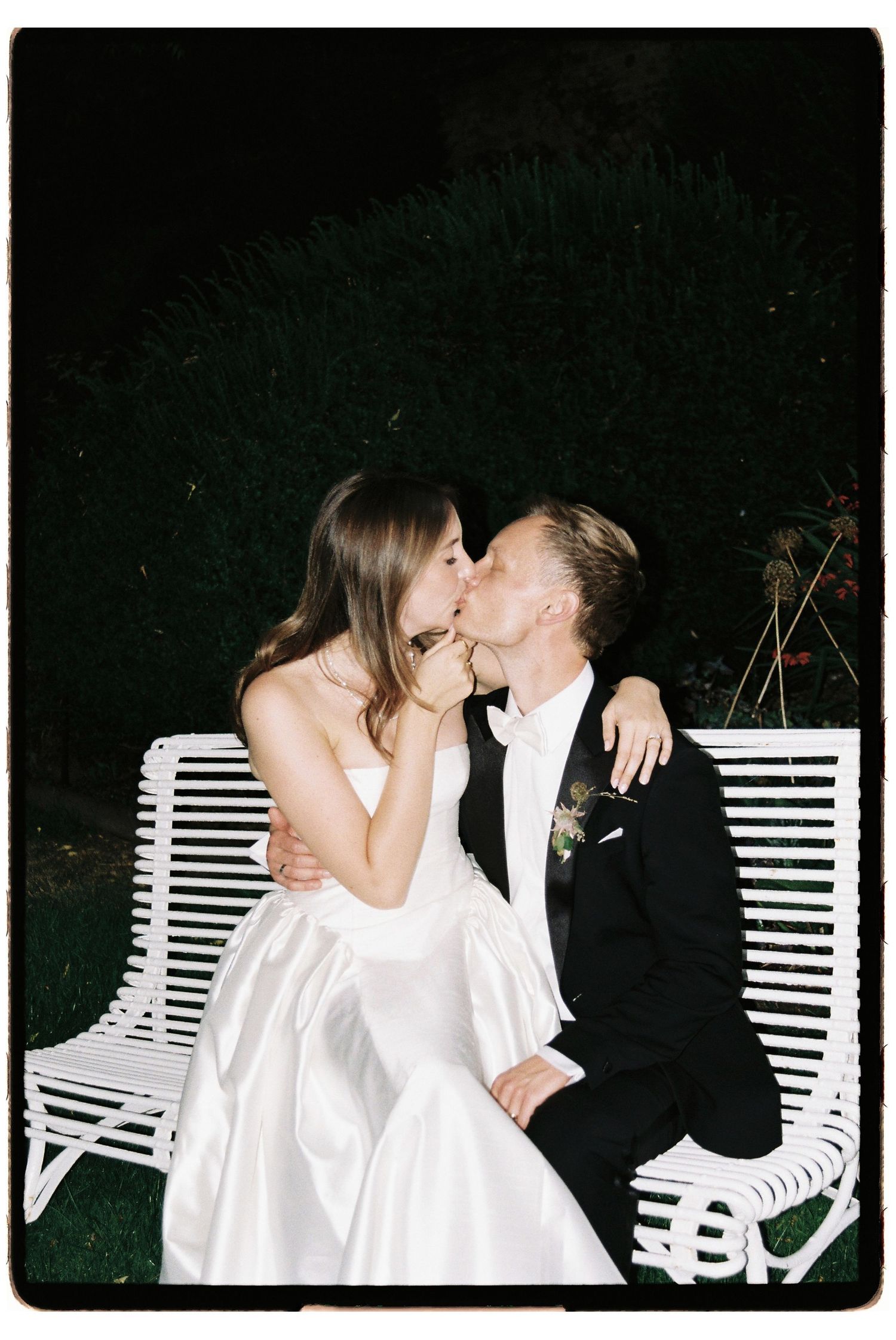 Bride and groom sharing a romantic kiss on a white bench outdoors at night during their wedding celebration.