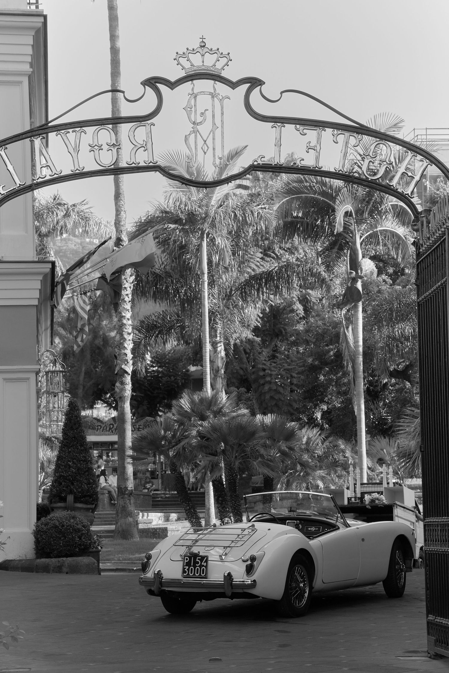 Vintage white convertible car parked under ornate Hotel Royal entrance arch, palm trees in black and white.