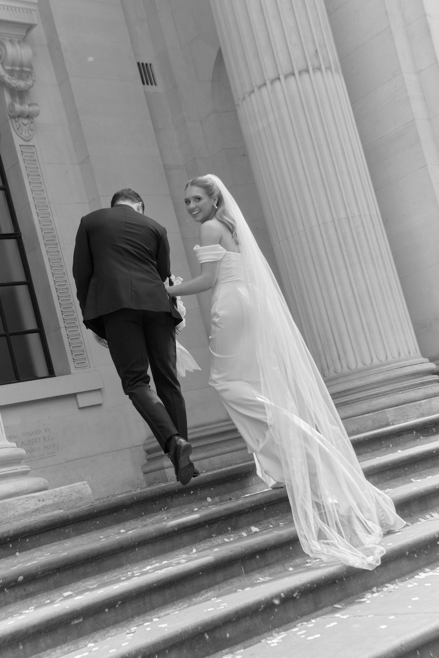 Black and white photo of a bride and groom walking up steps of a grand building with tall columns.