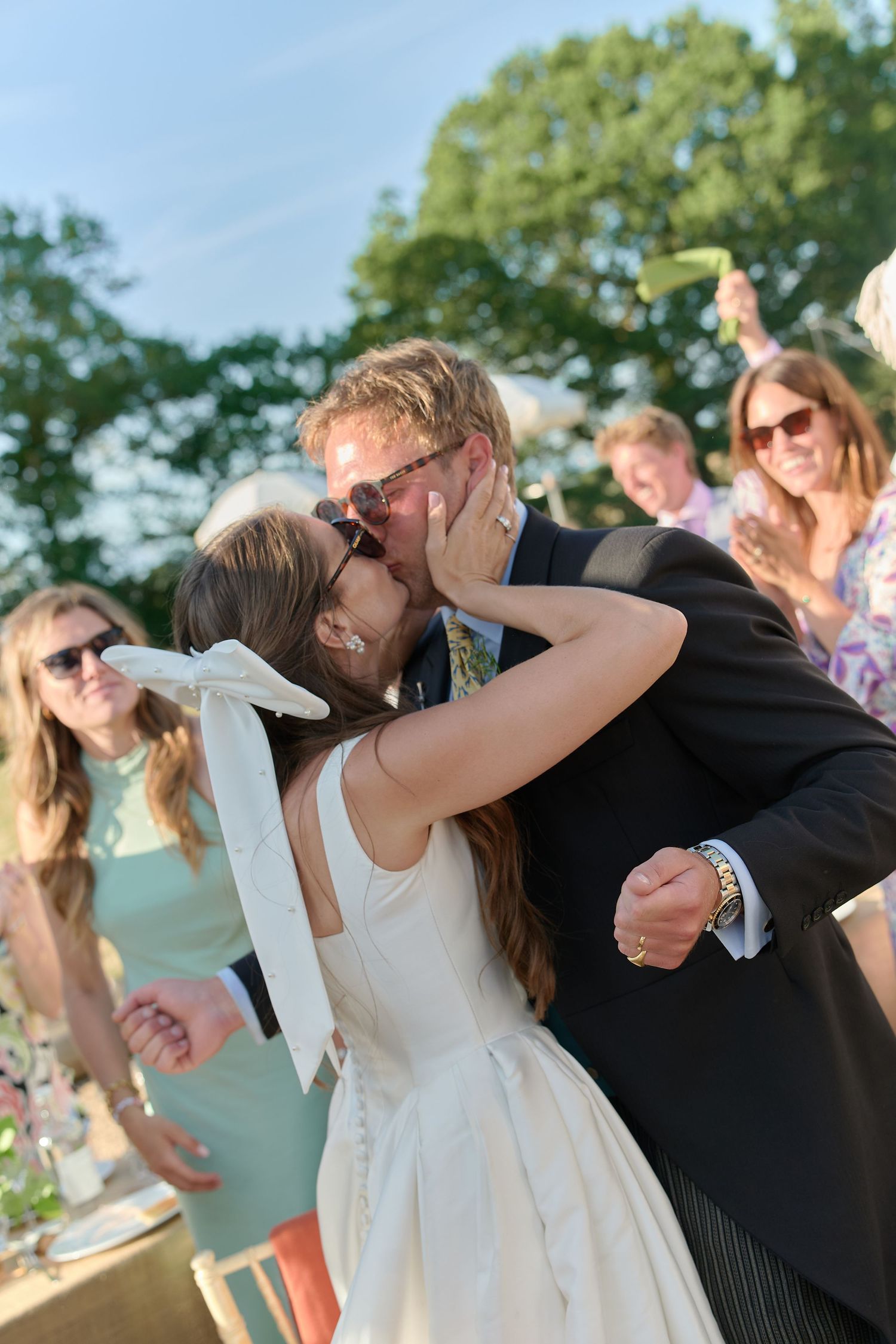 Bride and groom sharing a joyful kiss surrounded by celebrating wedding guests outdoors.