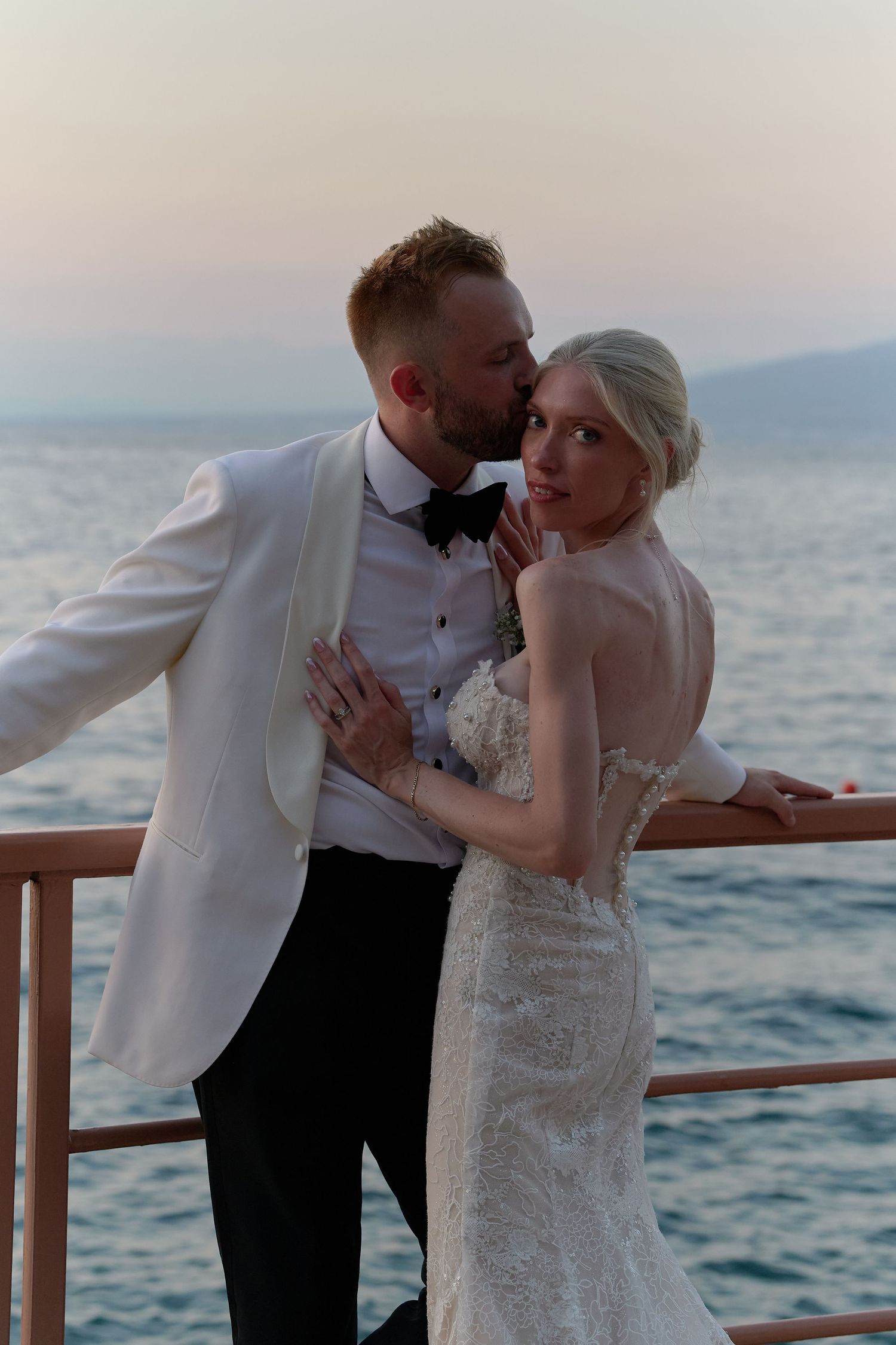 Bride and groom share a romantic kiss on a waterfront railing at sunset, she in lace gown, he in white tuxedo jacket.