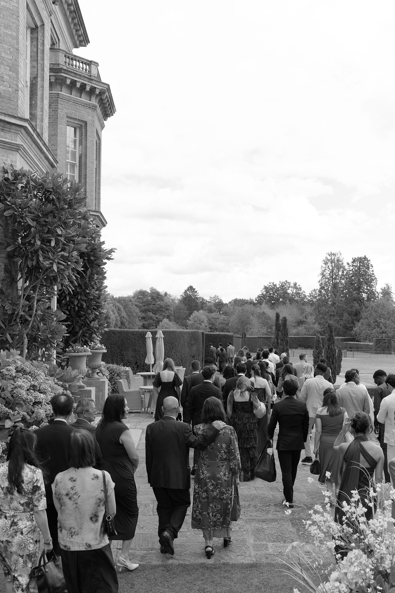 Guests walking toward an outdoor wedding ceremony at a grand estate, surrounded by lush gardens, in black and white.