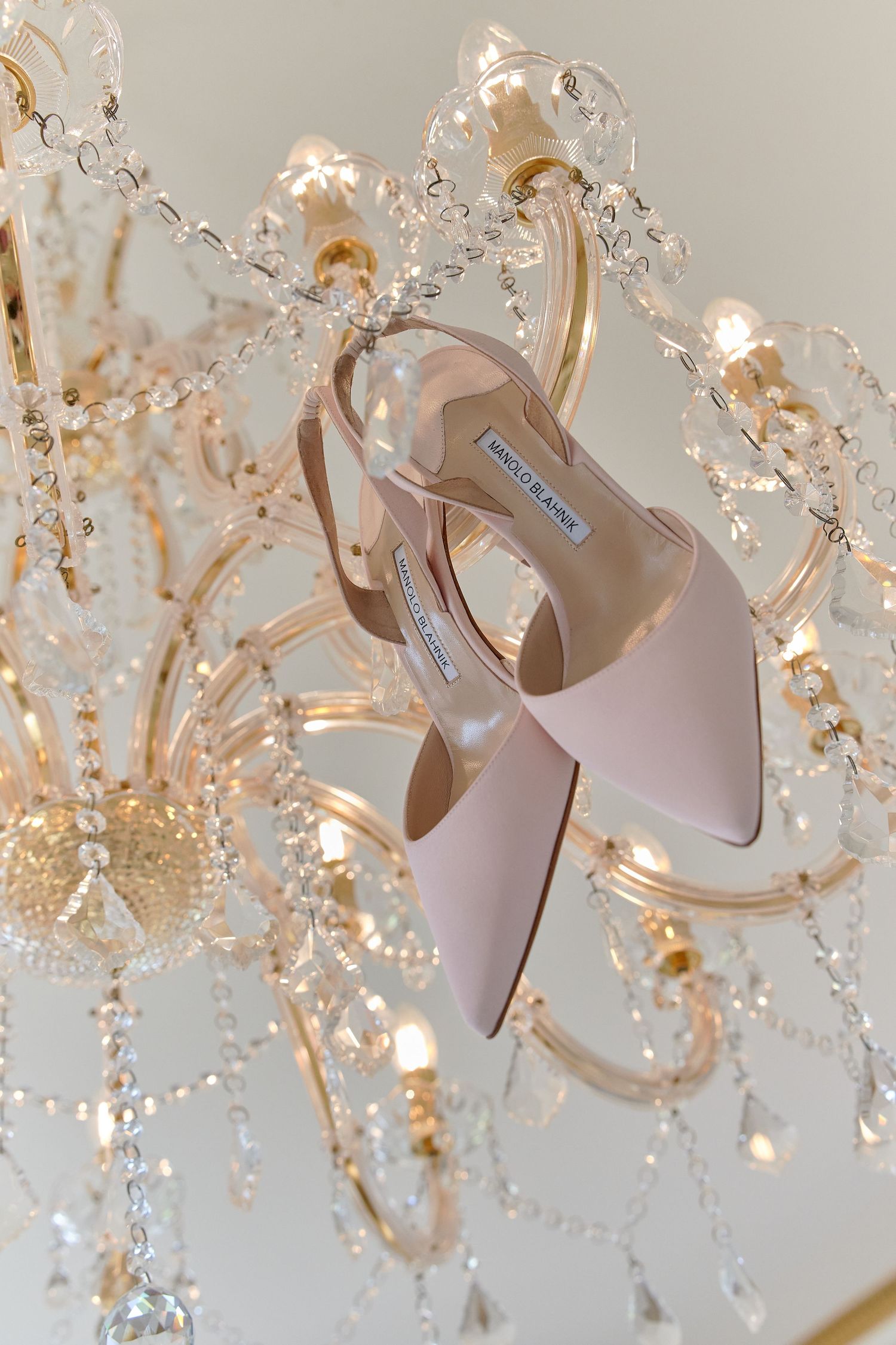 Blush pink Manolo Blahnik pointed-toe slingback heels hanging elegantly from a crystal chandelier.