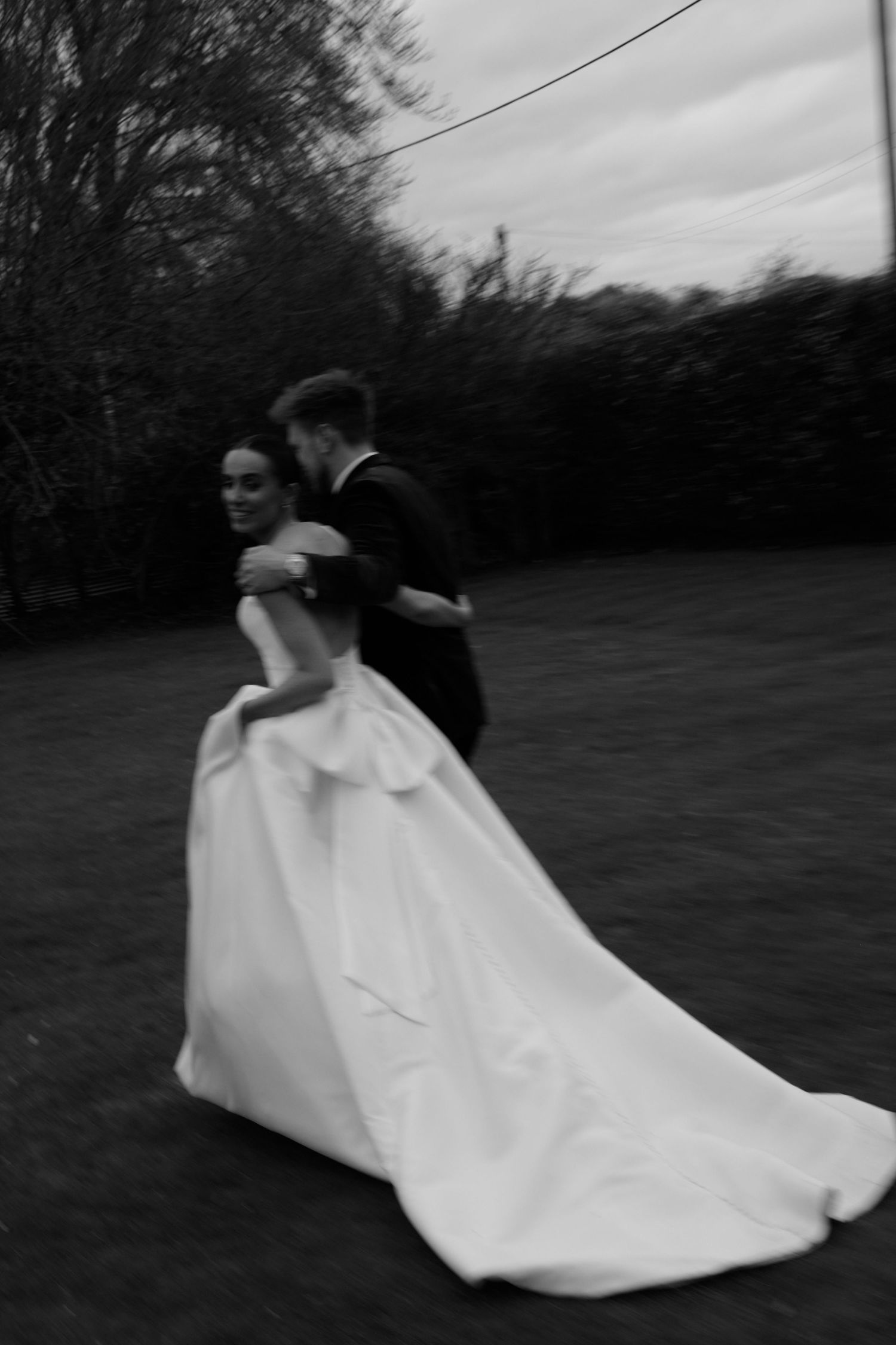 Black and white photo of a bride in a flowing gown and groom in a suit dancing outdoors on a lawn.