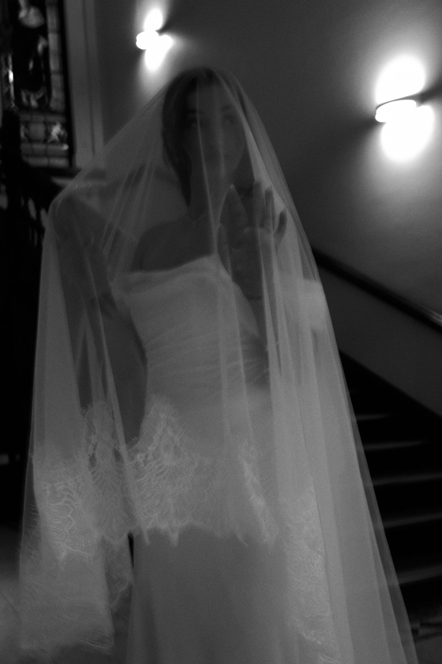 Bride in wedding gown and veil descending staircase in dramatic black and white photo.