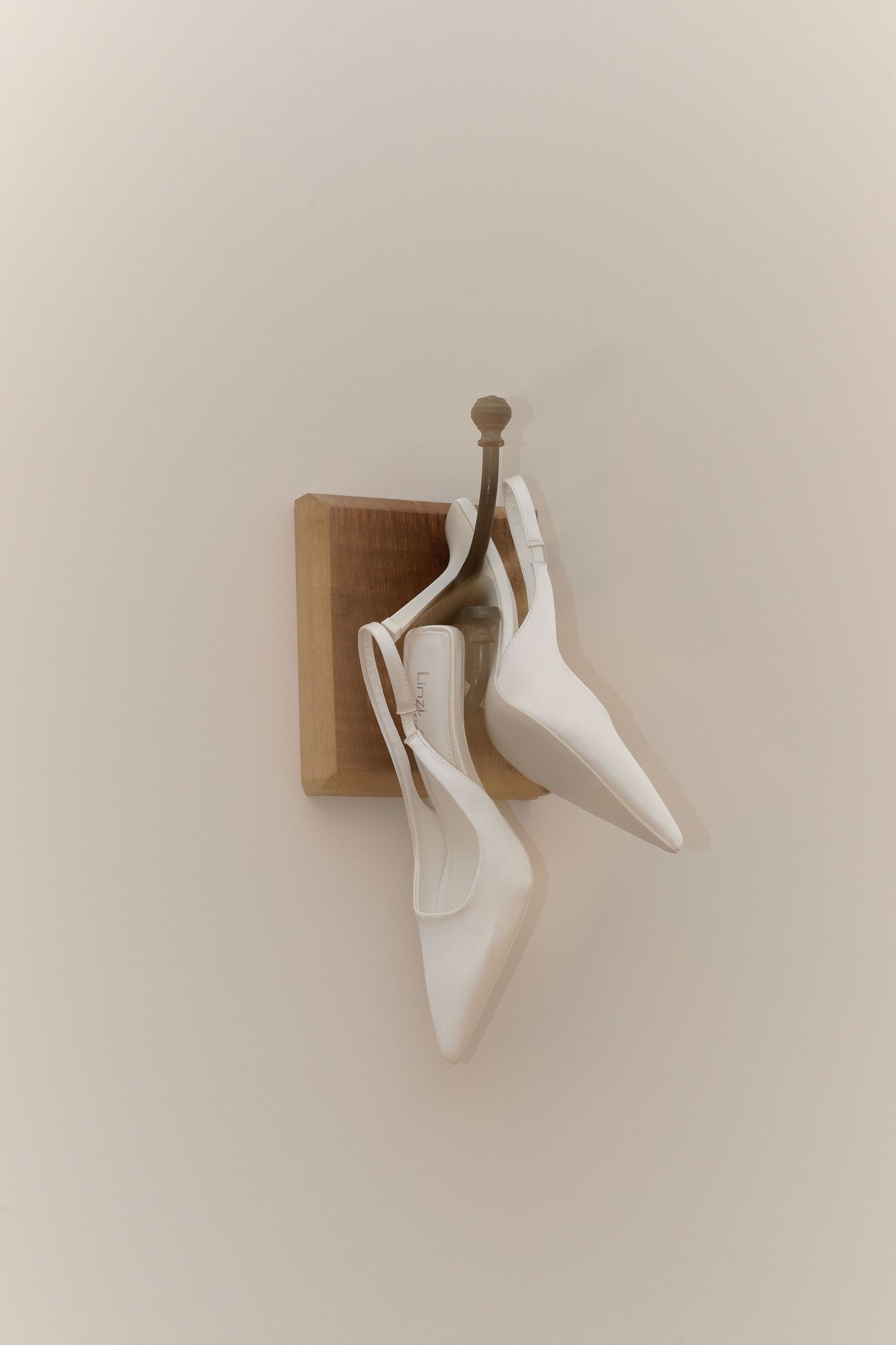 White slingback bridal heels hanging on wooden wall hook against neutral background.