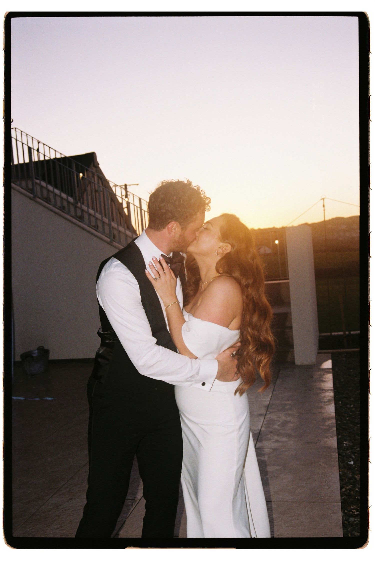 Newlyweds sharing romantic kiss on rooftop terrace during stunning golden hour sunset.