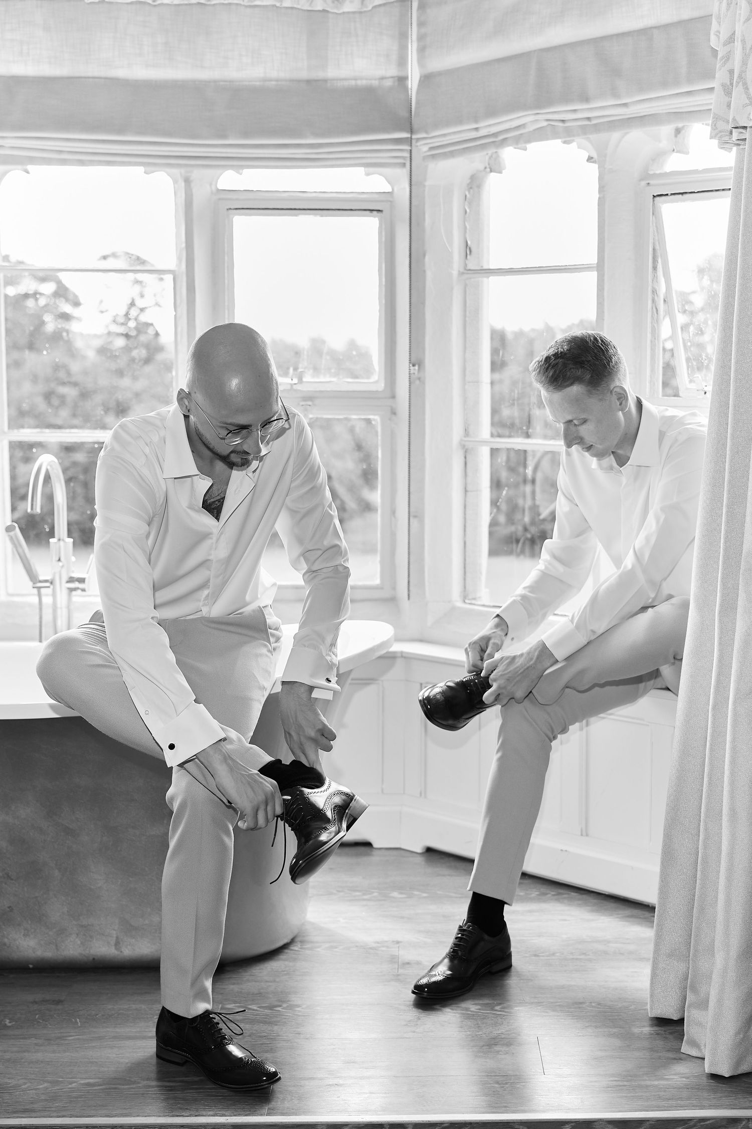 Two men in white dress shirts putting on formal black shoes while getting ready for a wedding ceremony.