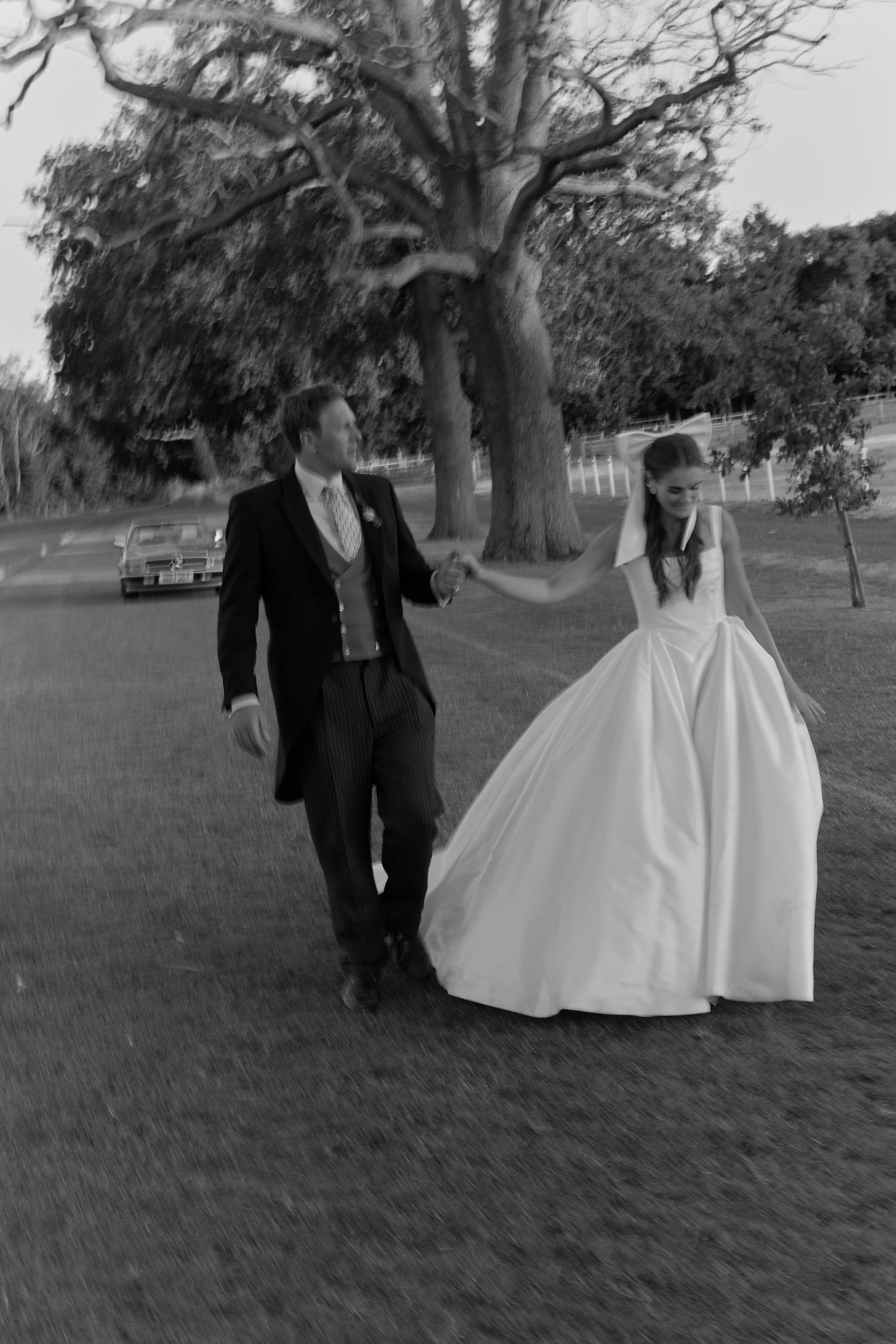 Black and white photo of bride and groom walking hand in hand under a large tree.