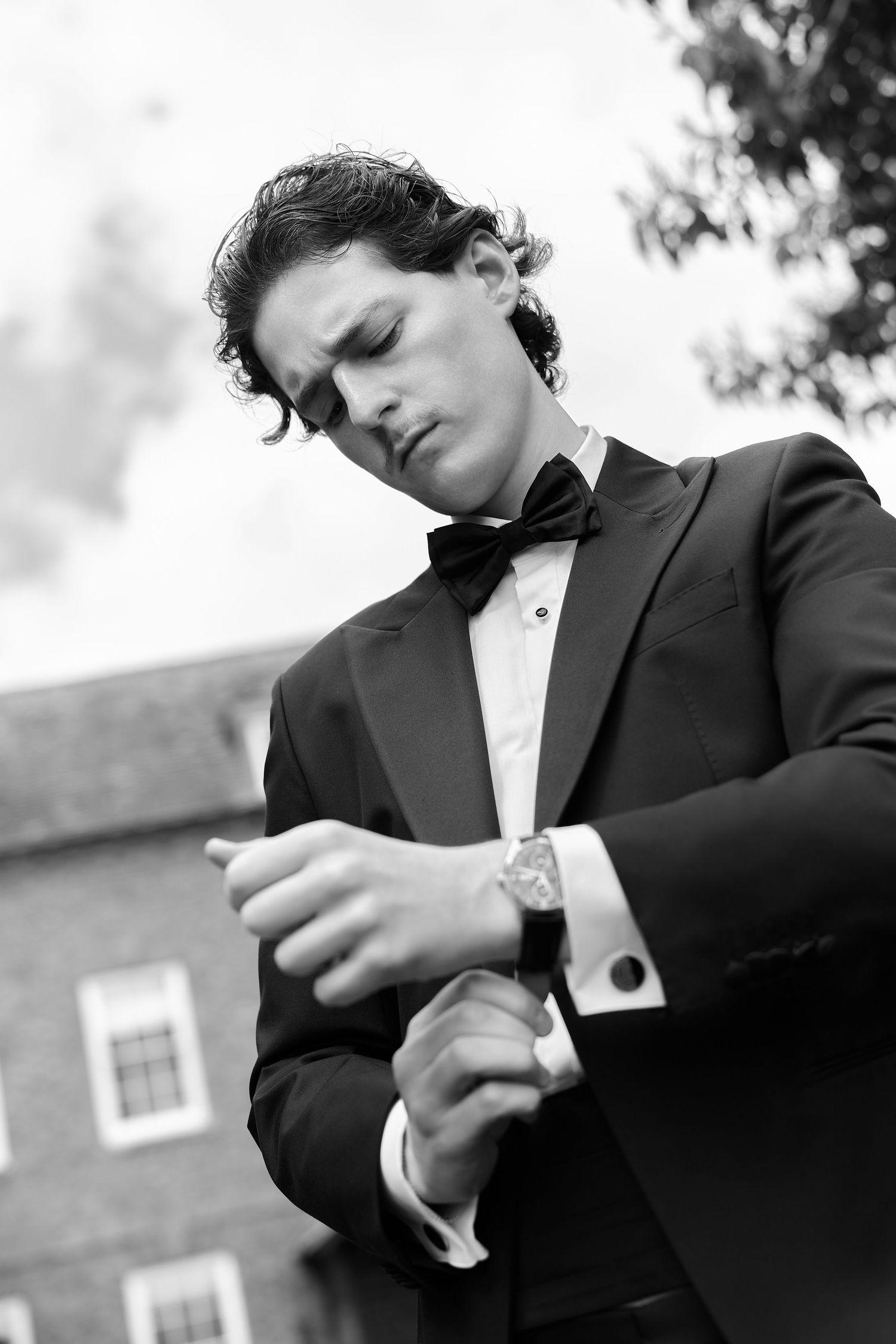 Elegant man in tuxedo and bow tie checking his watch outdoors in black and white photo.