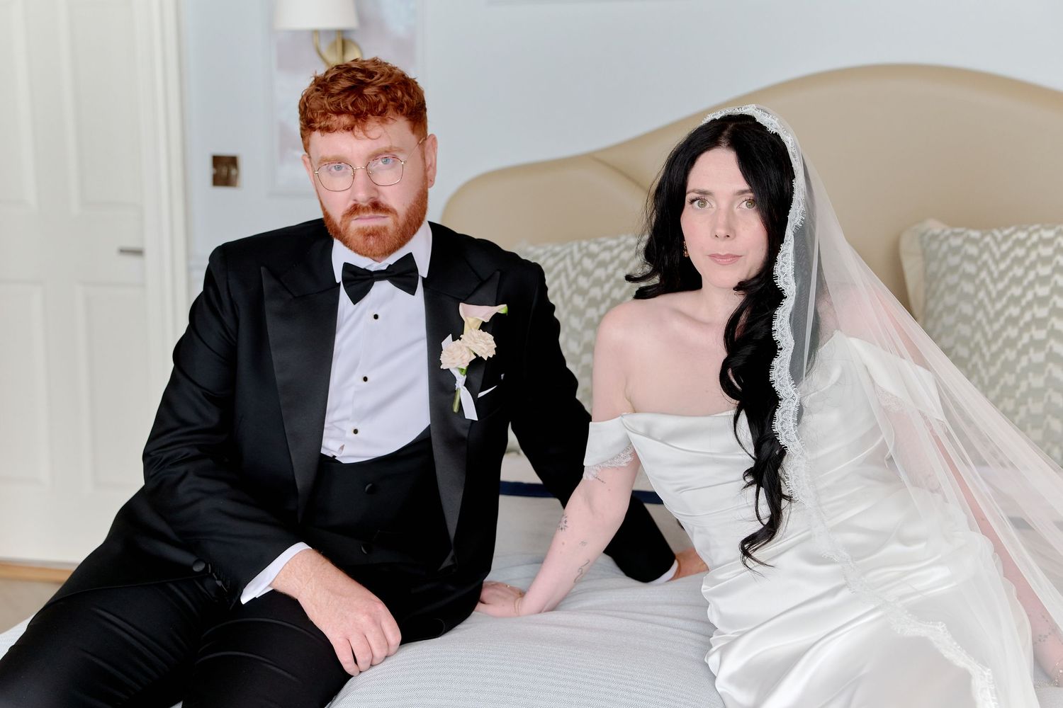 A bride in white gown and groom in black tuxedo sitting together on a bed in a formal wedding portrait.