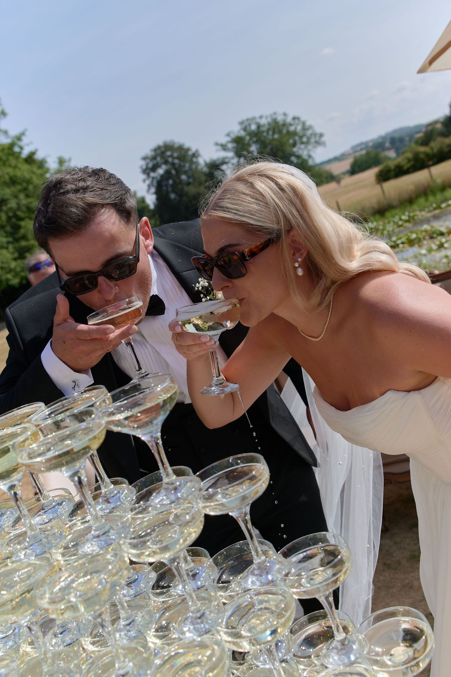 Bride and groom in sunglasses sipping champagne from a coupe tower at an outdoor wedding celebration.