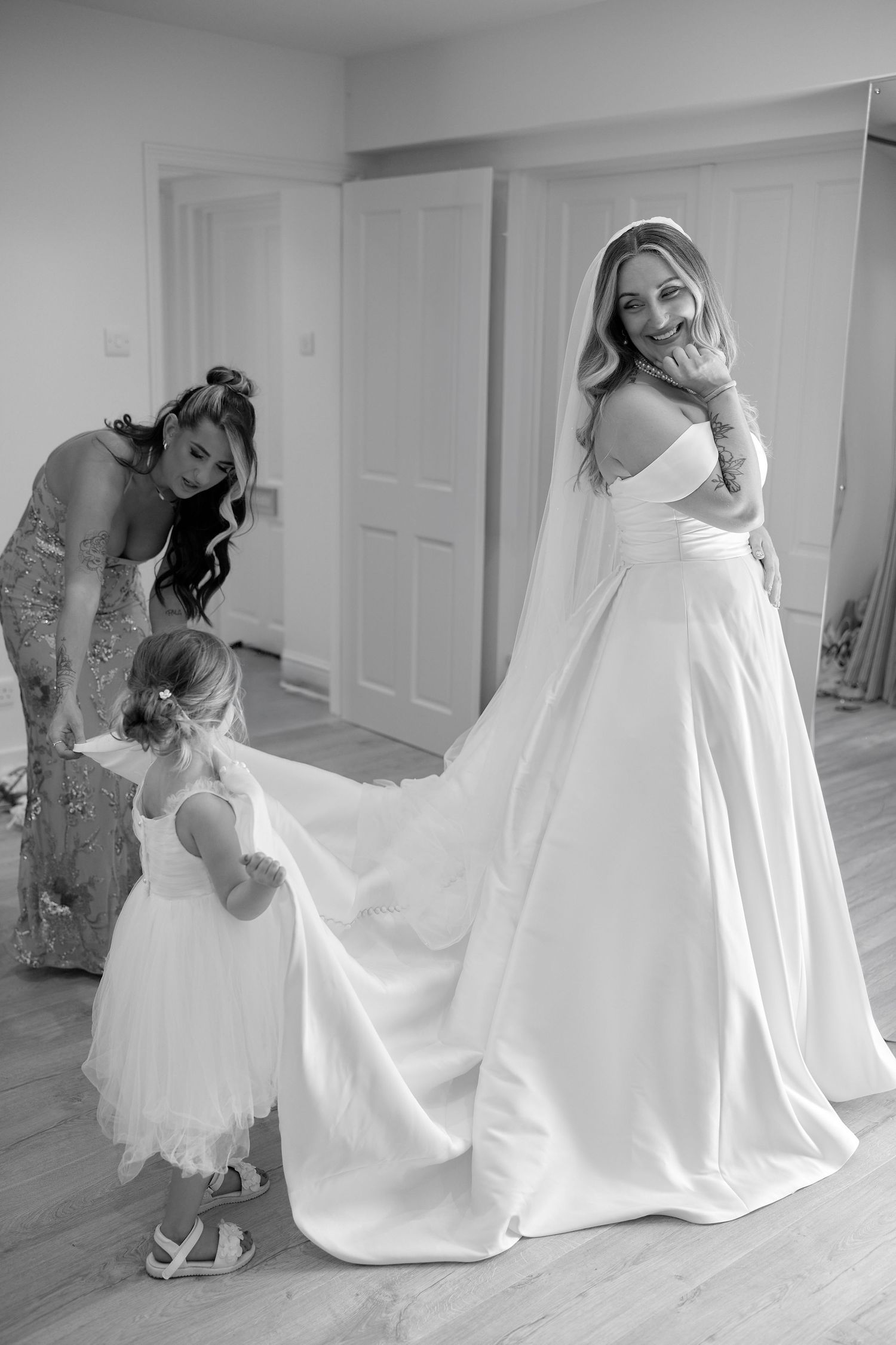 Black and white photo of a smiling bride in a white gown and veil, assisted by a bridesmaid and flower girl.