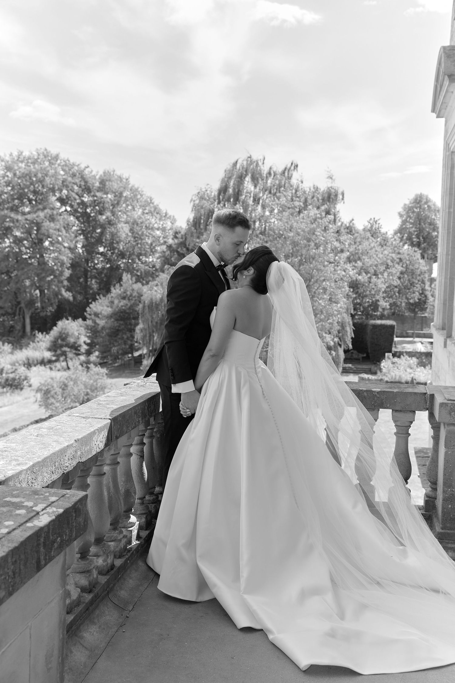 Black and white photo of a bride and groom sharing a kiss on a balcony, surrounded by lush trees on their wedding day.