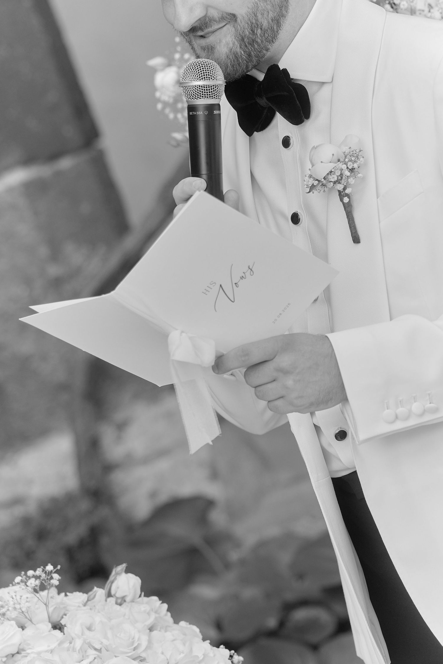Groom in white tuxedo reads wedding vows from paper card while holding microphone at outdoor ceremony.