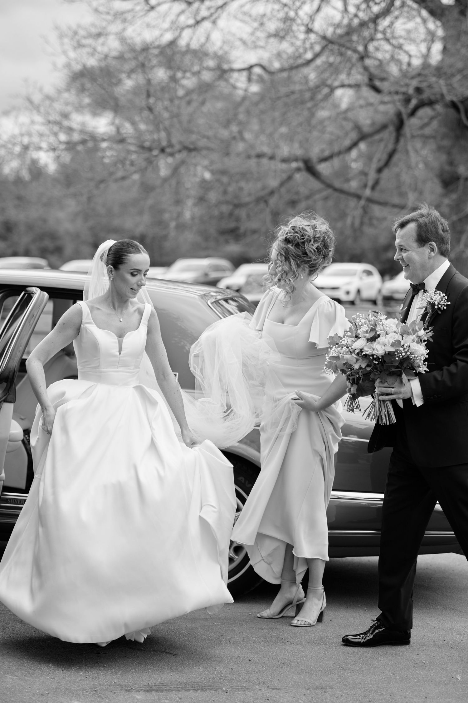 Bride exits car assisted by bridesmaid and groom holding bouquet, black and white wedding photo.