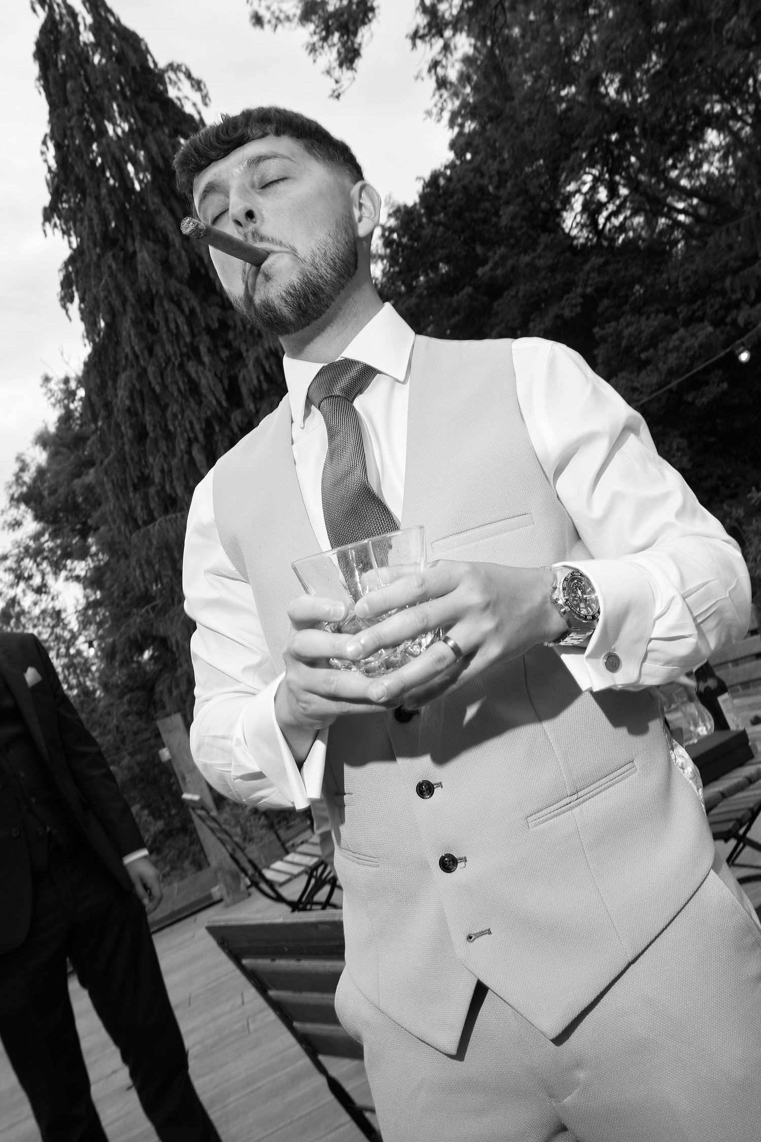 Man in white vest suit smoking cigar and holding whiskey glass outdoors in black and white photo.