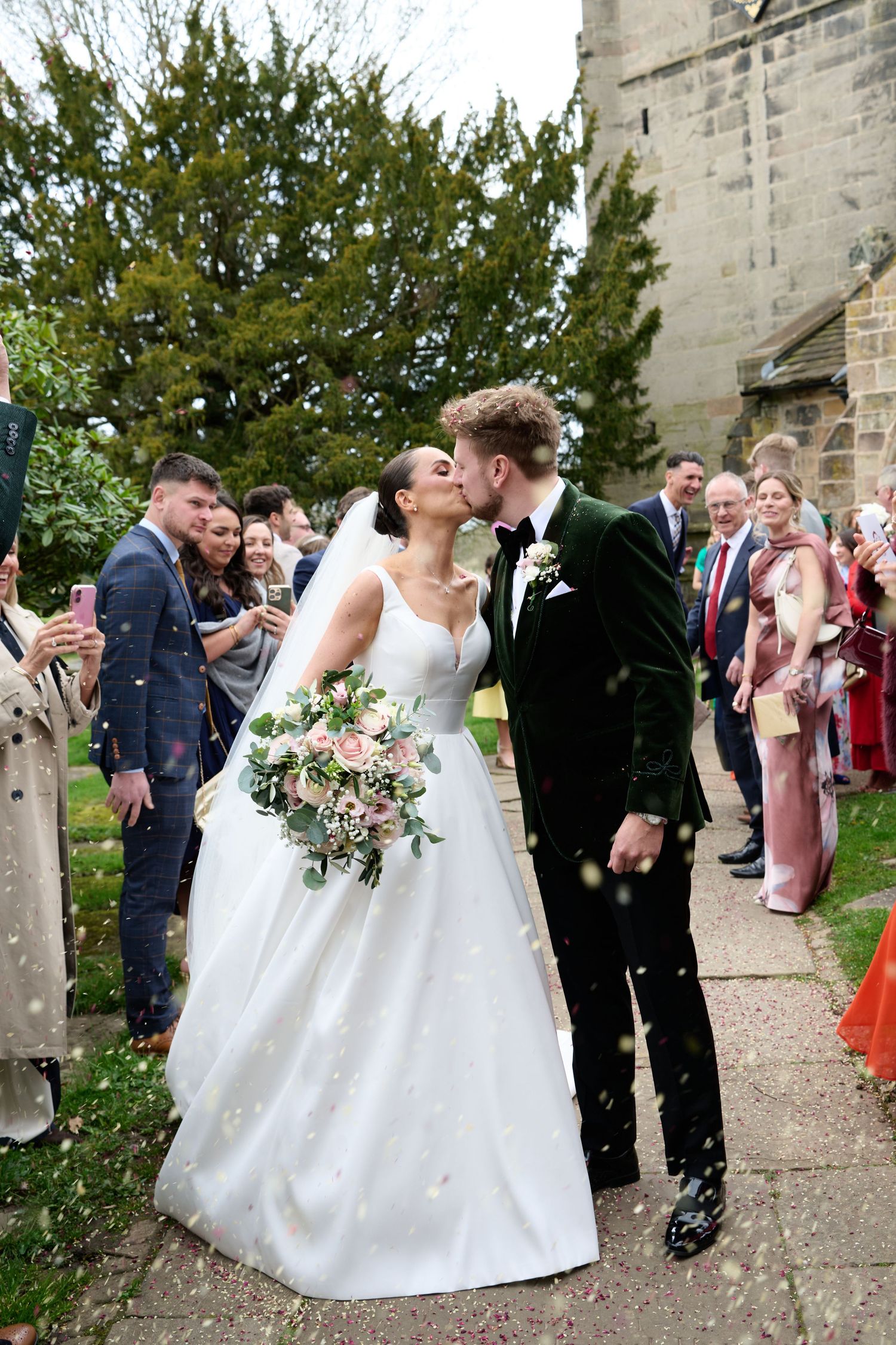 Bride and groom share a kiss while guests throw confetti outside a stone church on their wedding day.