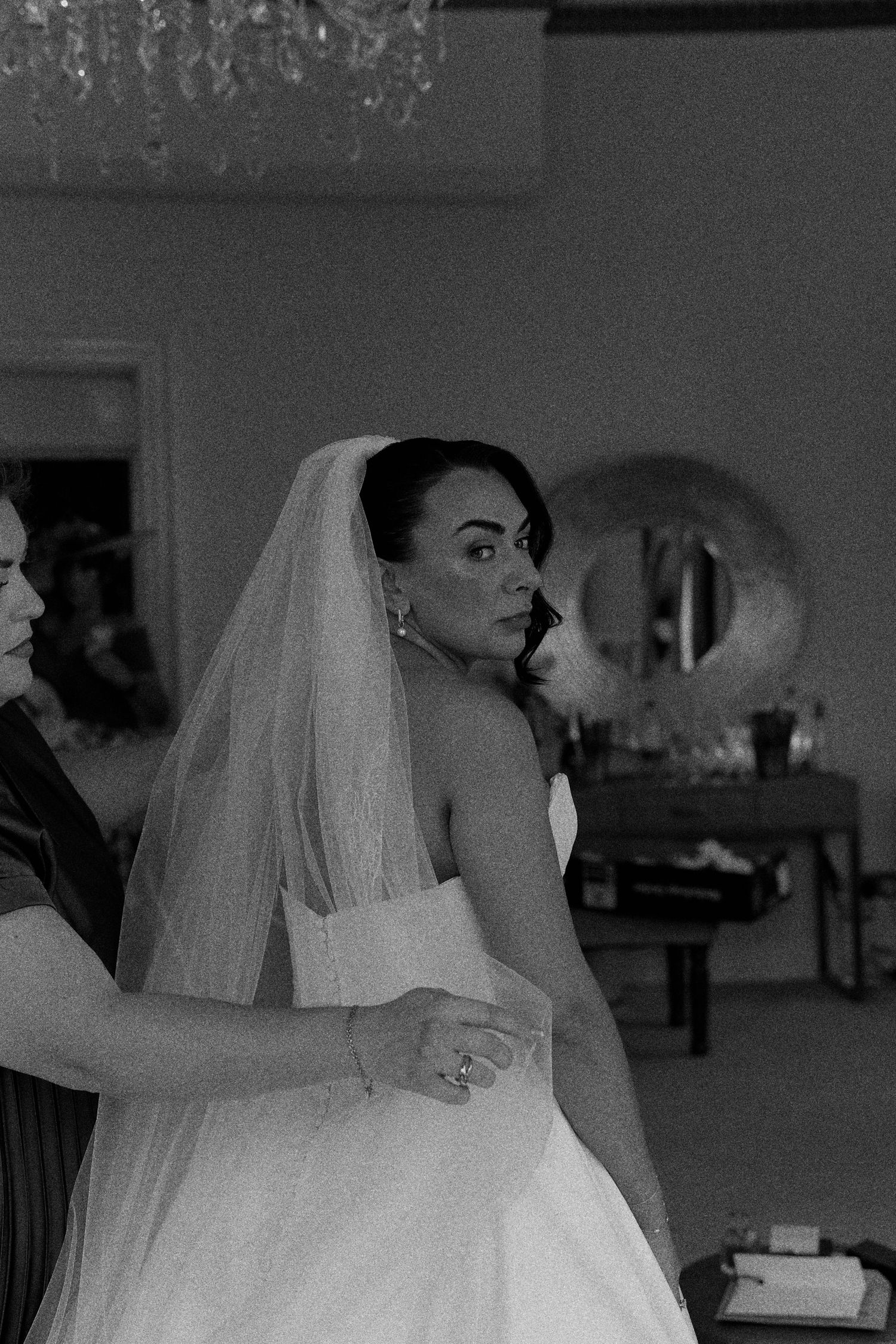 Black and white photo of a bride in a strapless gown and veil being helped by a woman in a chandelier-lit room.