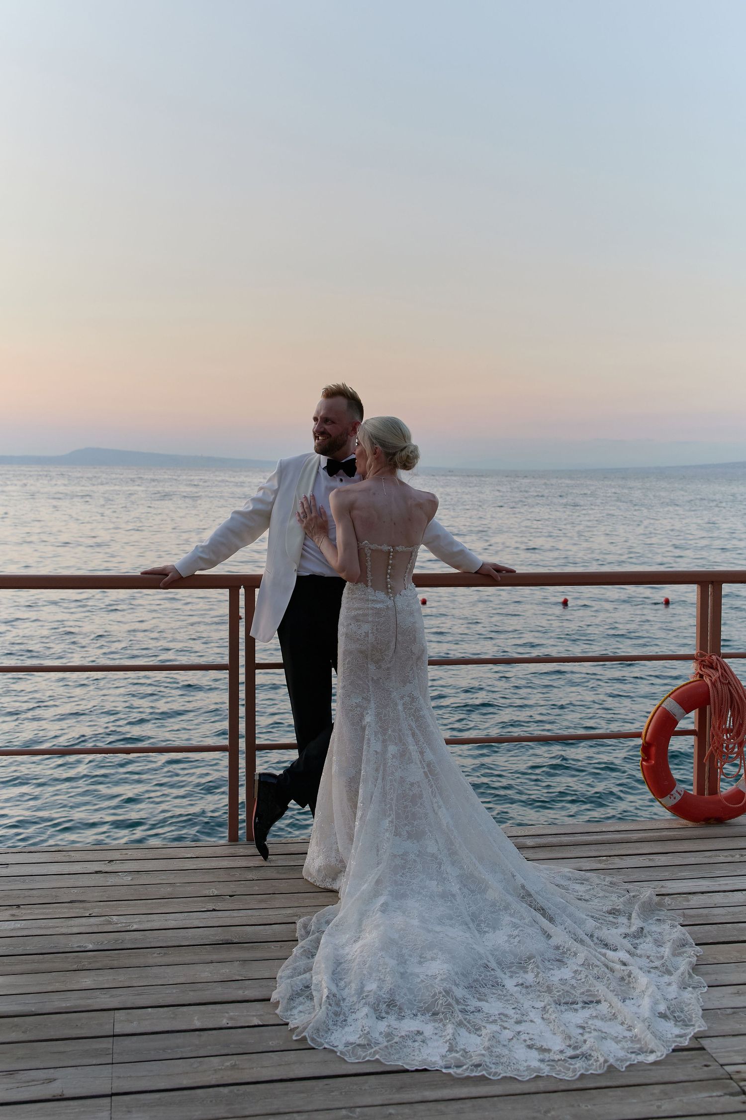 Bride and groom pose on a wooden pier at sunset, overlooking calm ocean waters, bride in lace gown, groom in white tuxedo.