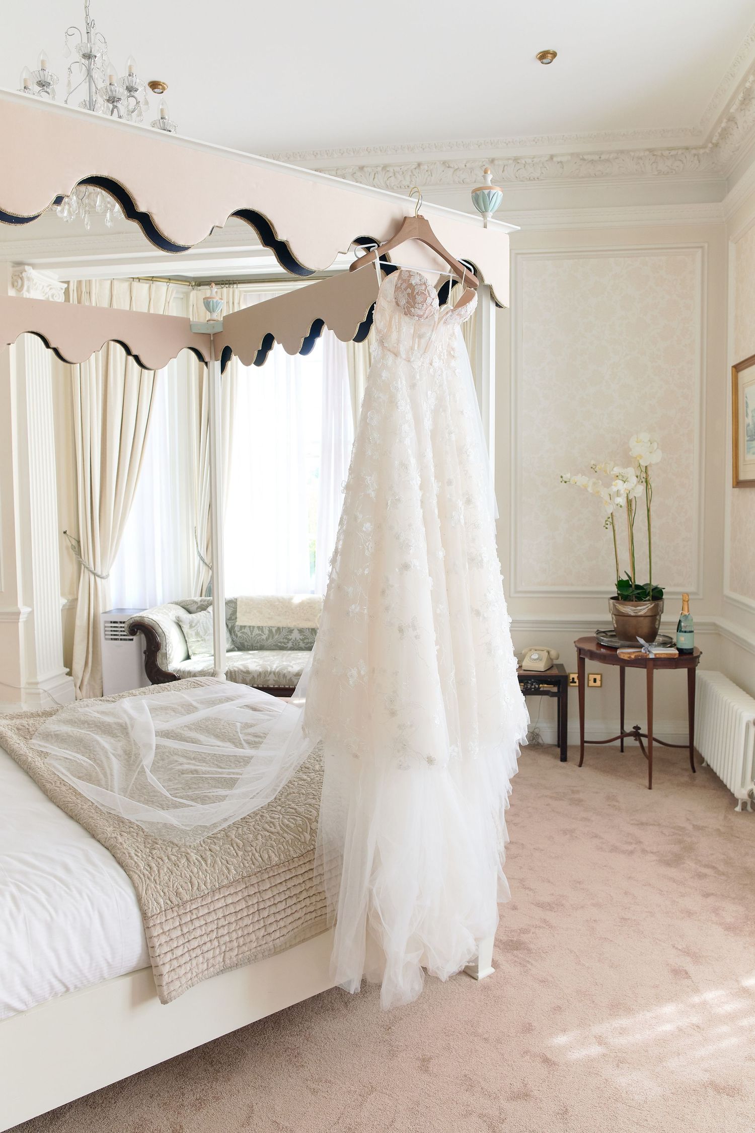 Elegant white wedding dress hanging on a four-poster bed frame in a luxurious cream bridal suite.