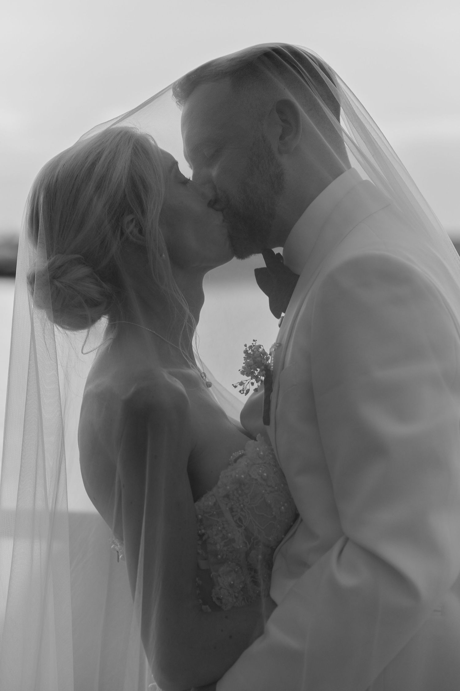 Black and white photo of bride and groom sharing a romantic kiss under a flowing veil on their wedding day.