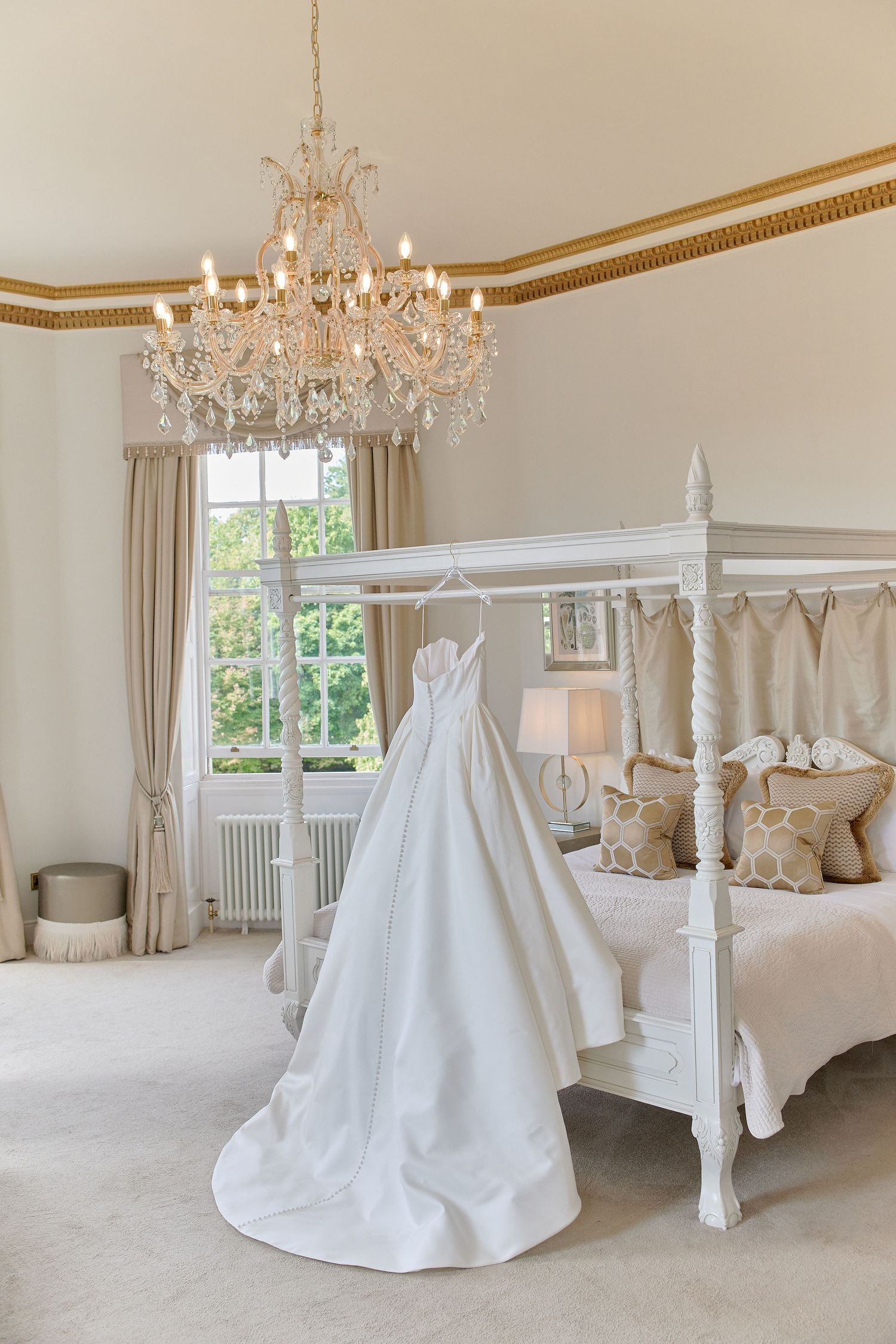 White wedding dress hanging on four-poster bed in luxurious bridal suite with crystal chandelier.