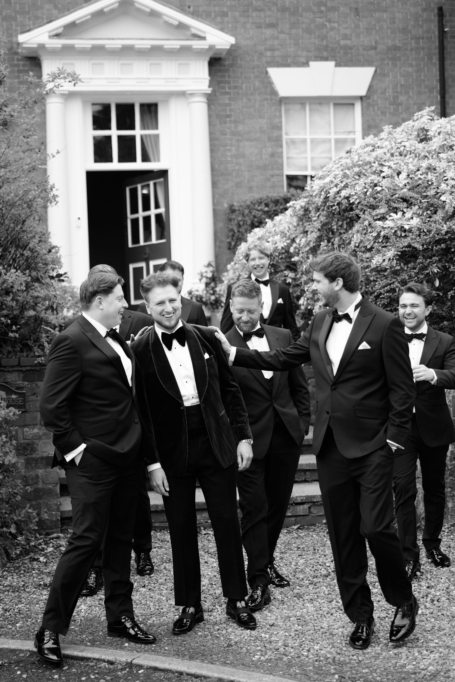Group of groomsmen in black tuxedos laughing outside an elegant brick building on a wedding day.