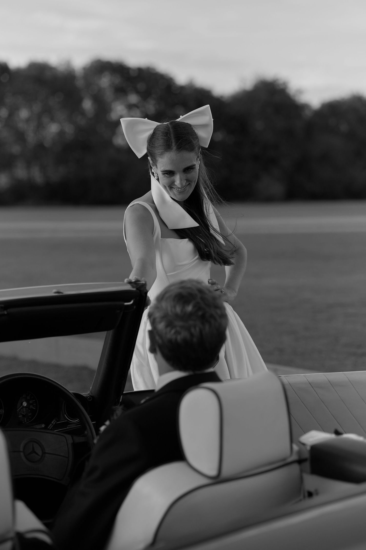 Black and white photo of a smiling bride with a bow veil leaning into a convertible.