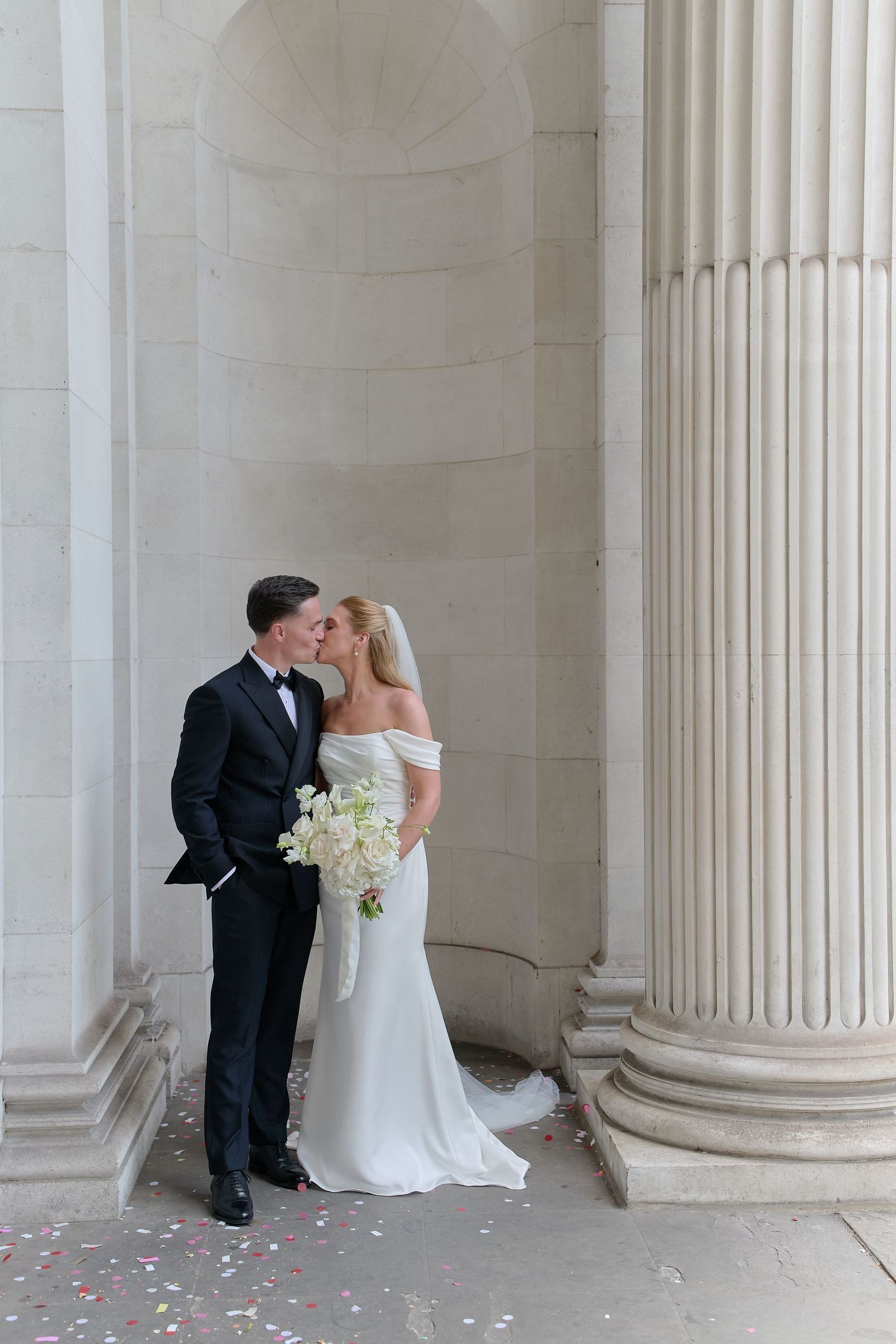 Bride and groom share a kiss beside elegant stone columns, bride holding white bouquet with confetti on the ground.