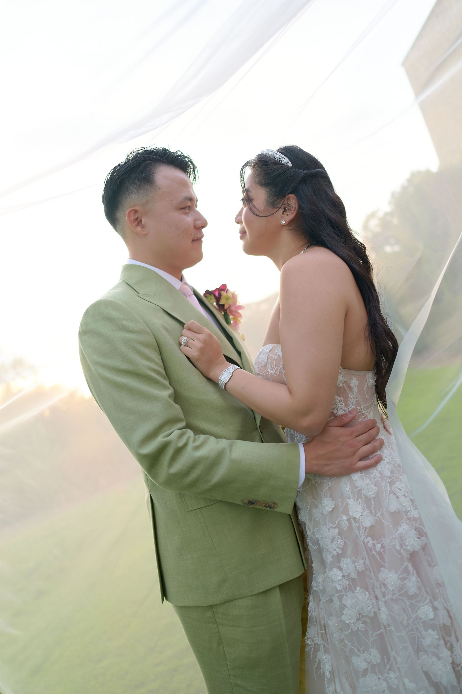 Bride and groom in sage suit share romantic moment under sheer drape at outdoor wedding ceremony.