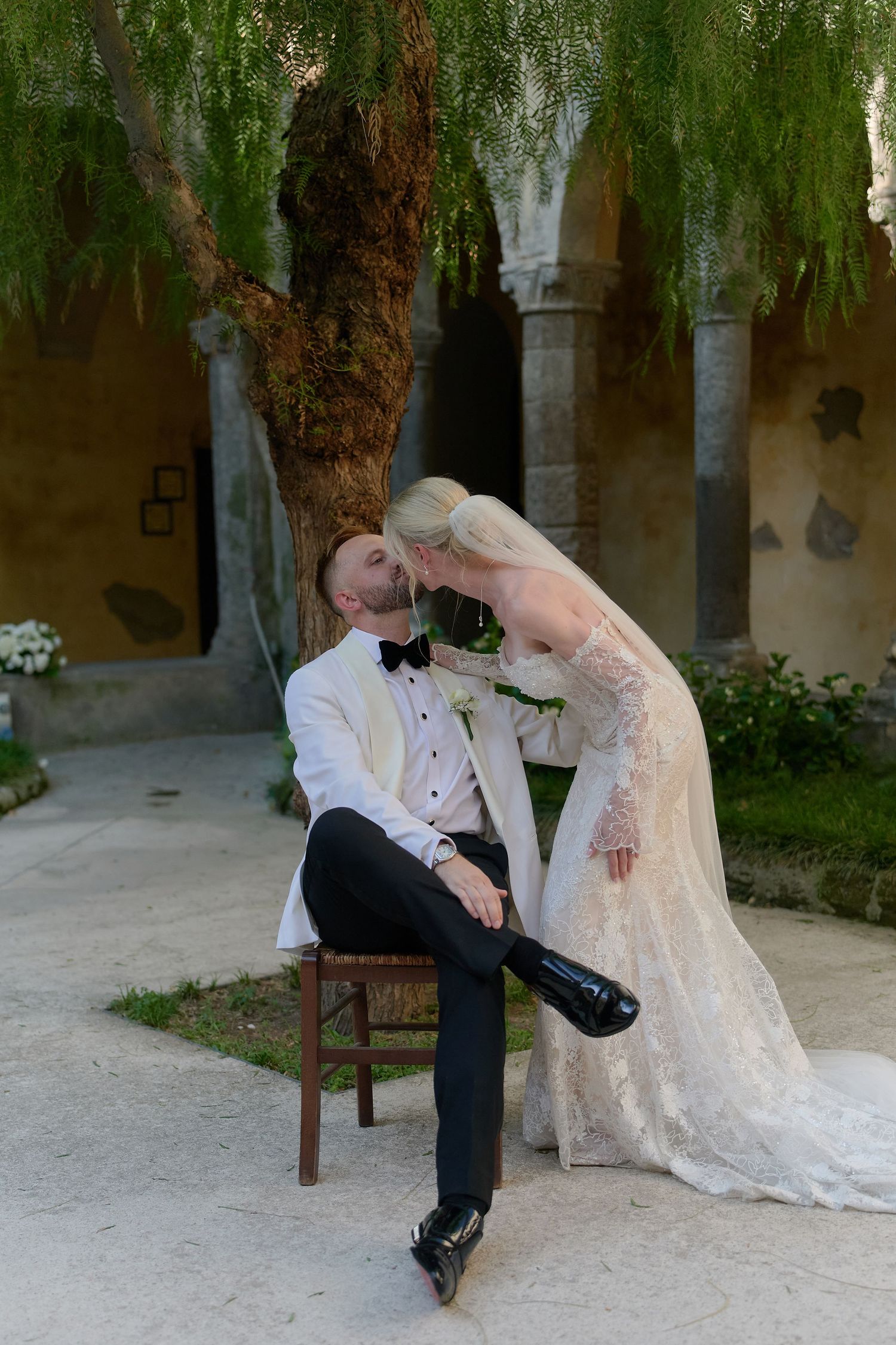 Bride in lace gown leans to kiss groom in white tuxedo seated on chair in elegant garden courtyard.