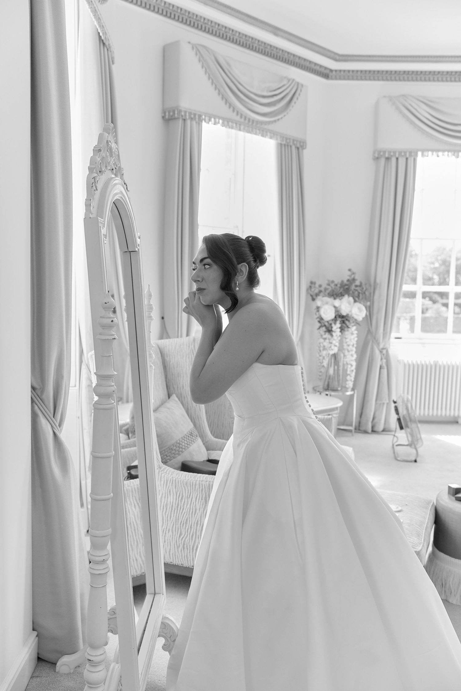 Bride in a strapless ball gown gazes into an ornate mirror in an elegant room, black and white wedding photo.