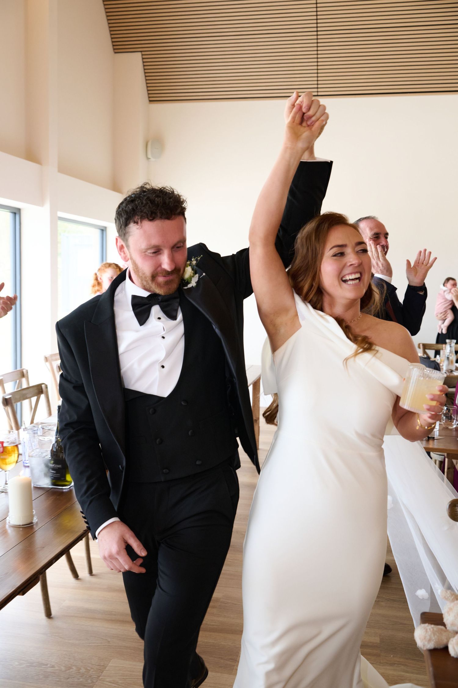 Joyful bride and groom make grand entrance at wedding reception, bride raising hand in celebration wearing white gown.