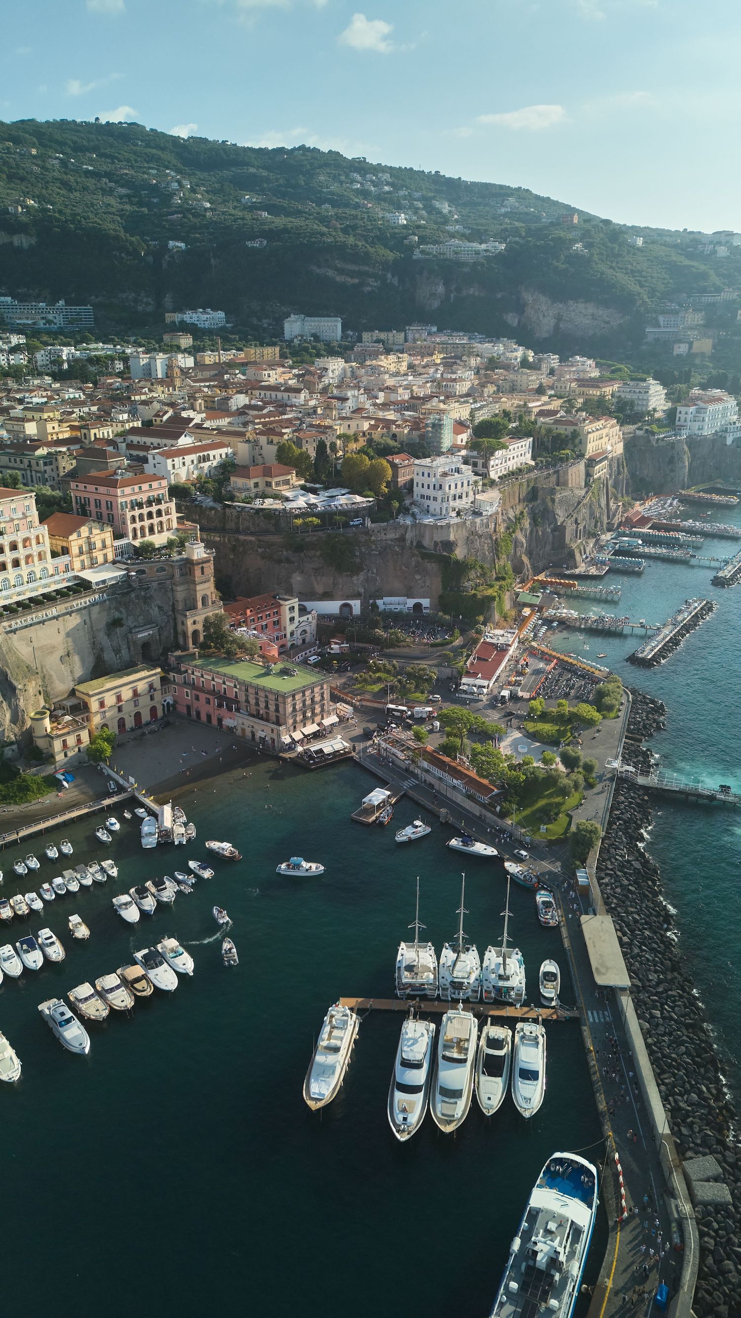 Aerial view of Sorrento Italy coastline with colorful buildings marina filled with white boats and lush green hillside backdrop.