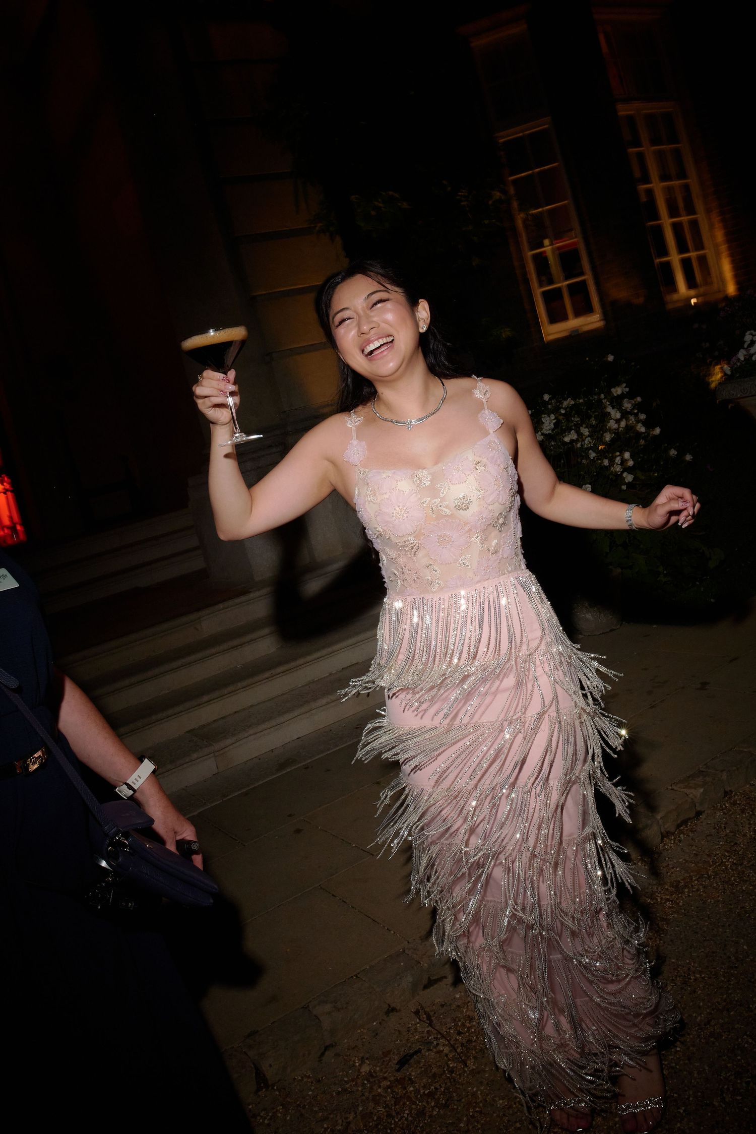 Woman in pink feathered sequin gown laughing and dancing outdoors at night, holding a champagne glass.