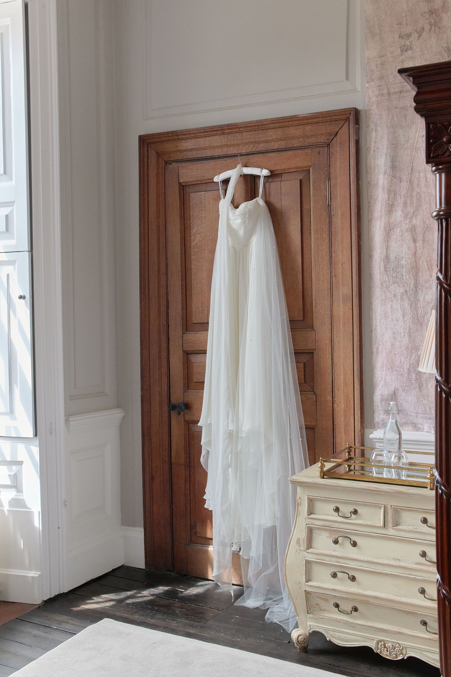 White wedding dress hanging on wooden door beside vintage dresser in elegant bridal suite.