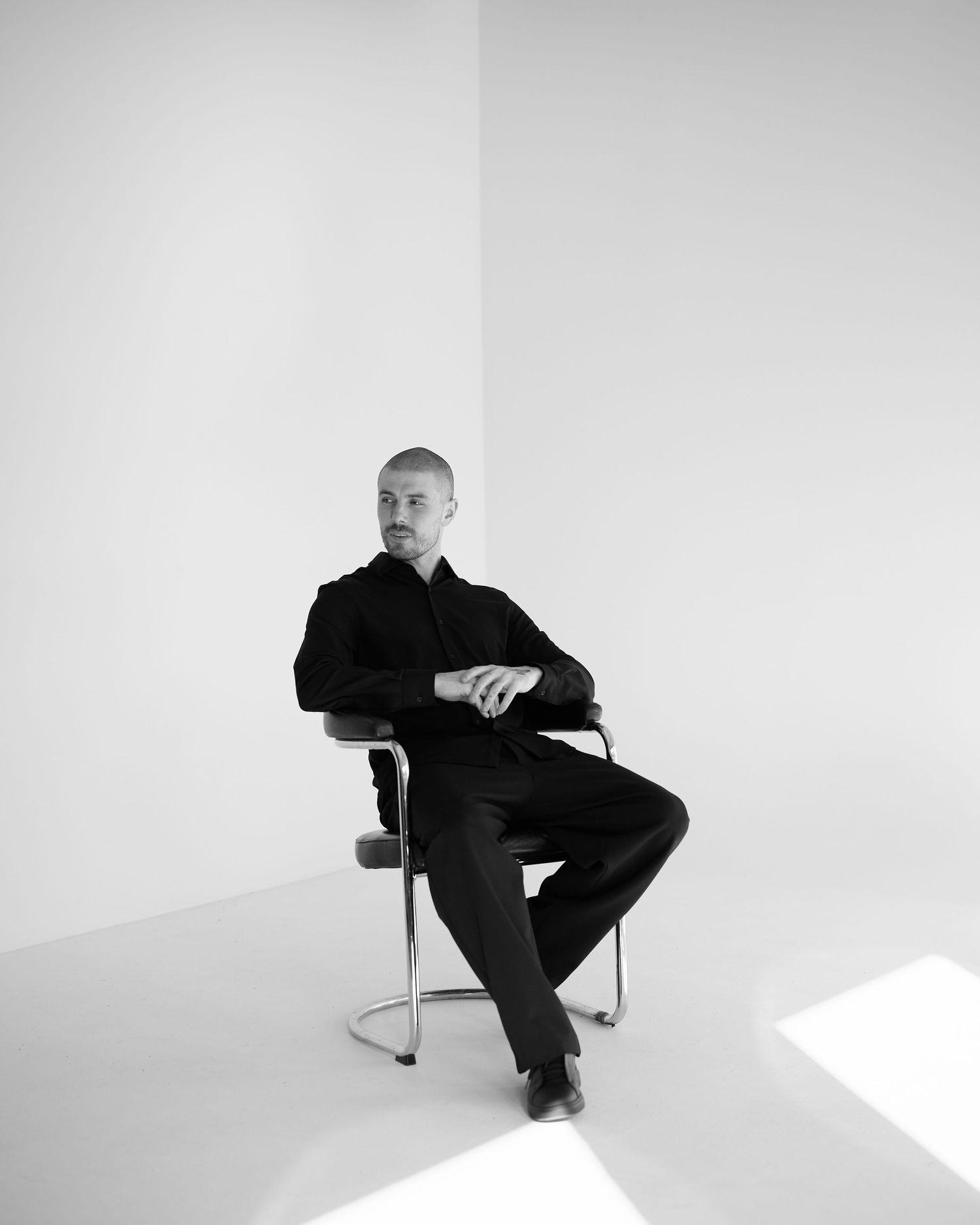 Man in black suit seated on chair in minimalist black and white studio portrait.